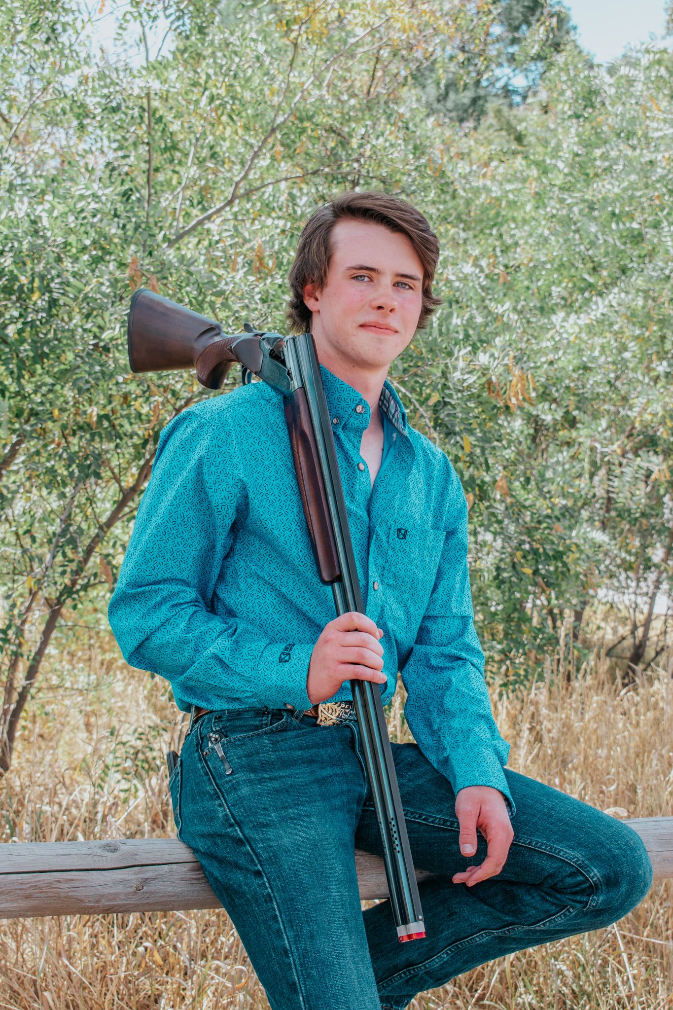 Young man in teal western shirt holding a shotgun while leaning on a wooden fence outdoors for senior photos.