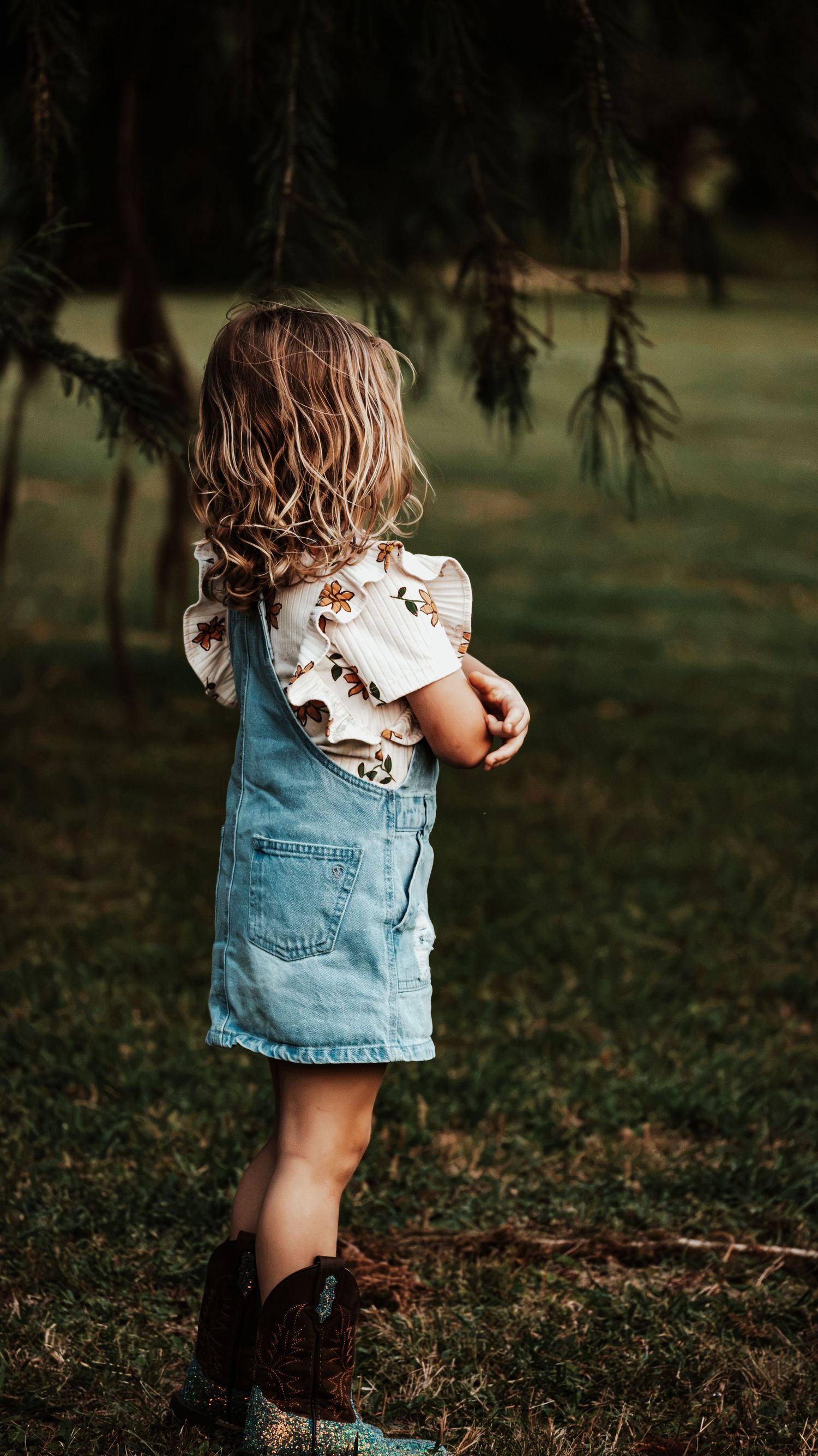 Toddler girl wearing denim overall dress and brown cowboy boots stands outdoors on grass.
