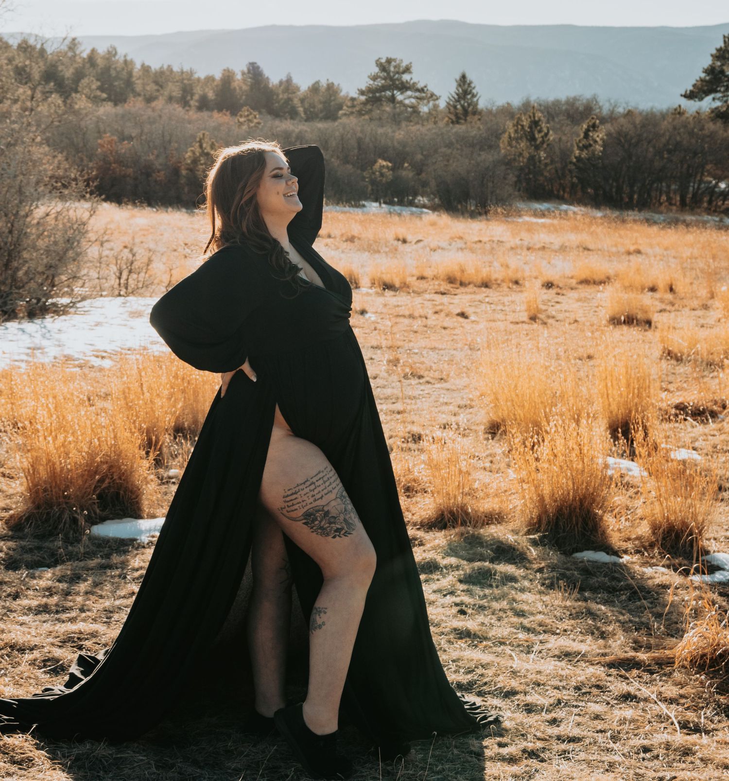 Pregnant woman in dramatic black maternity gown poses in golden winter field with snowy backdrop.