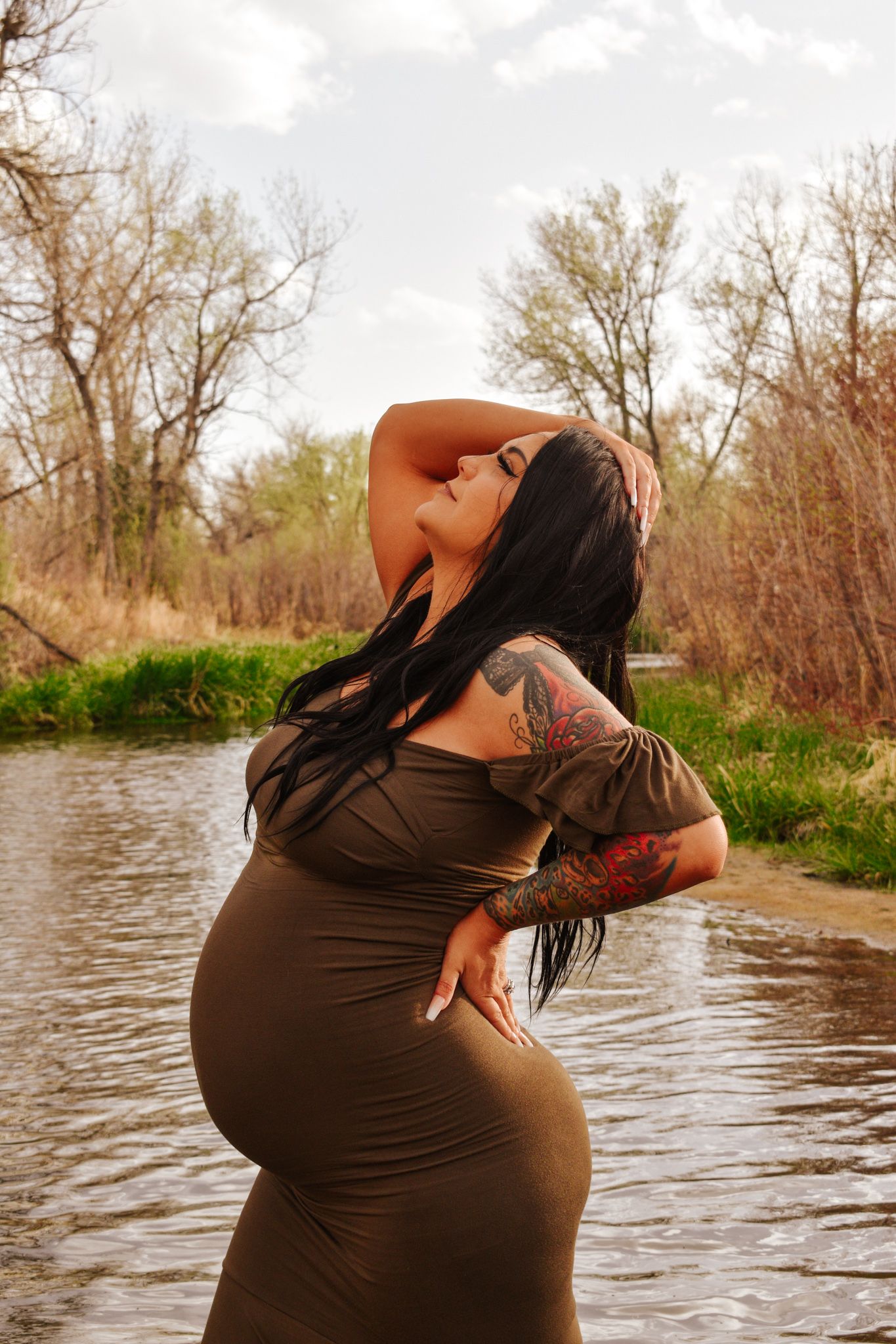Tattooed pregnant woman in olive dress posing by stream with hand raised in her hair.