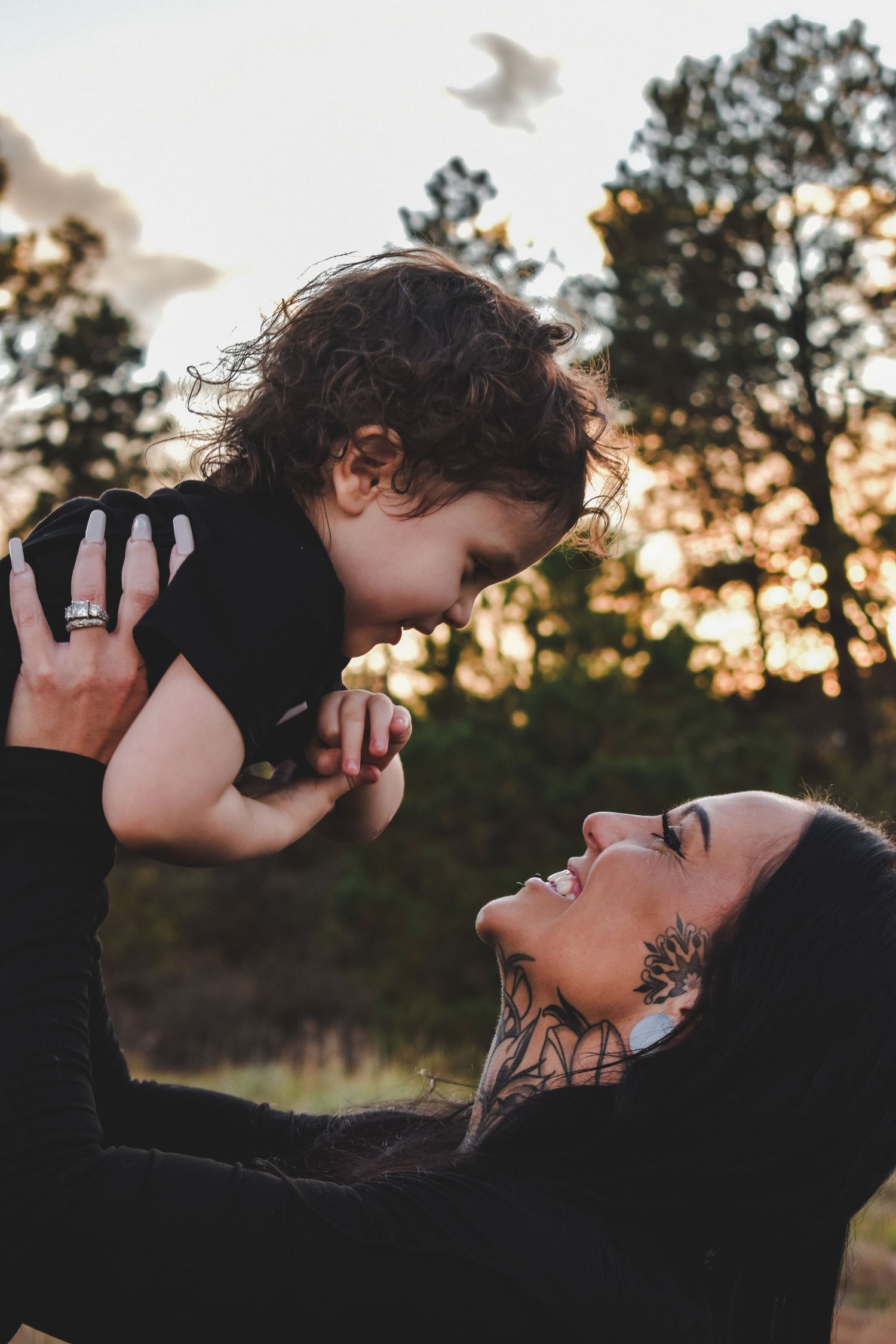 A tattooed mother joyfully lifts her curly-haired toddler against a golden sunset sky with pine trees in background.