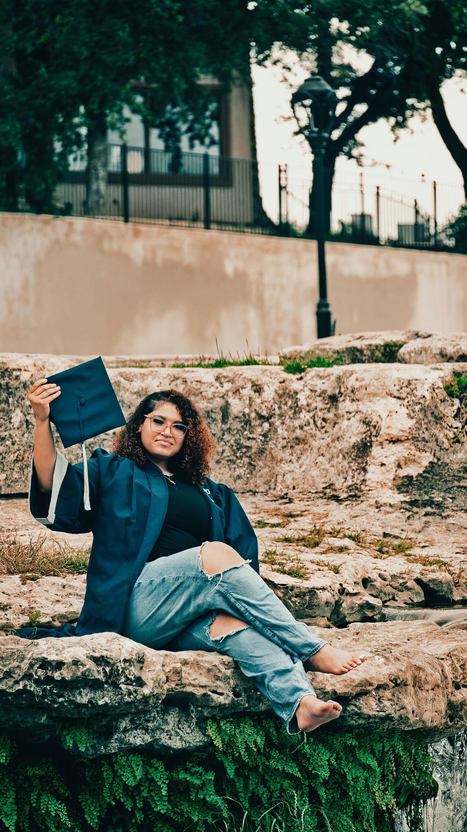 Graduate in blue cap and gown sitting on stone ruins holding mortarboard with trees behind for senior photos.