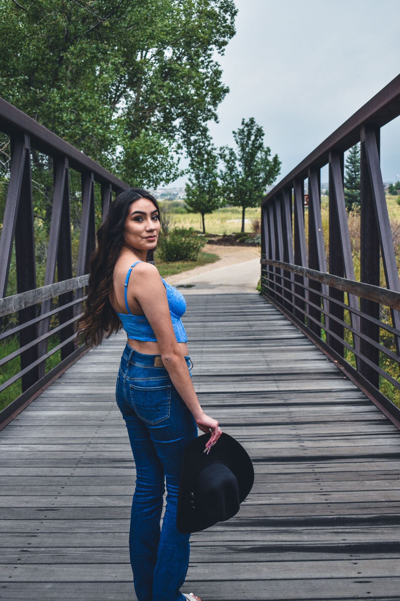 Young woman in blue crop top and jeans holds black hat while standing on a rustic wooden bridge for senior photos.