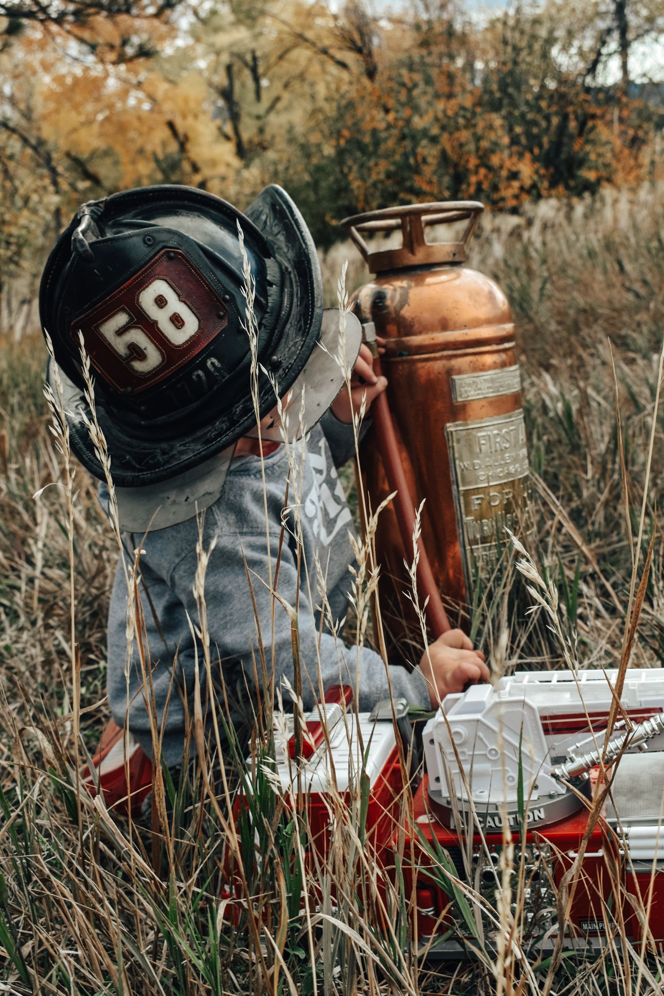 Toddler wearing firefighter helmet number 58 sitting with vintage copper fire extinguisher and toy fire truck in field.
