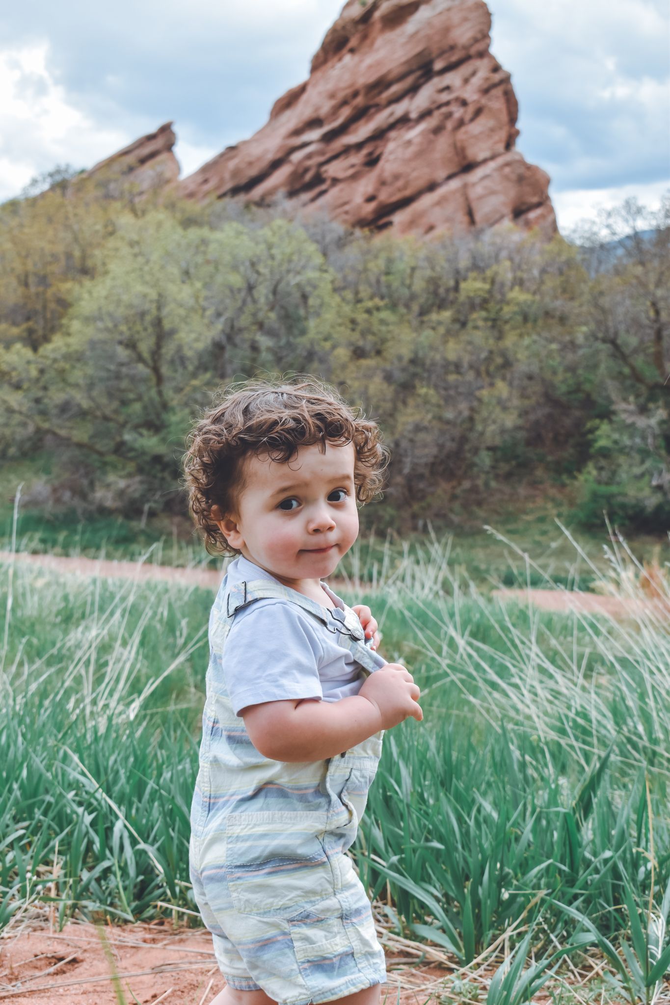 Curly-haired toddler stands in green meadow with red rock formations and trees in background. Family Photographer in Pflugerville, Tx