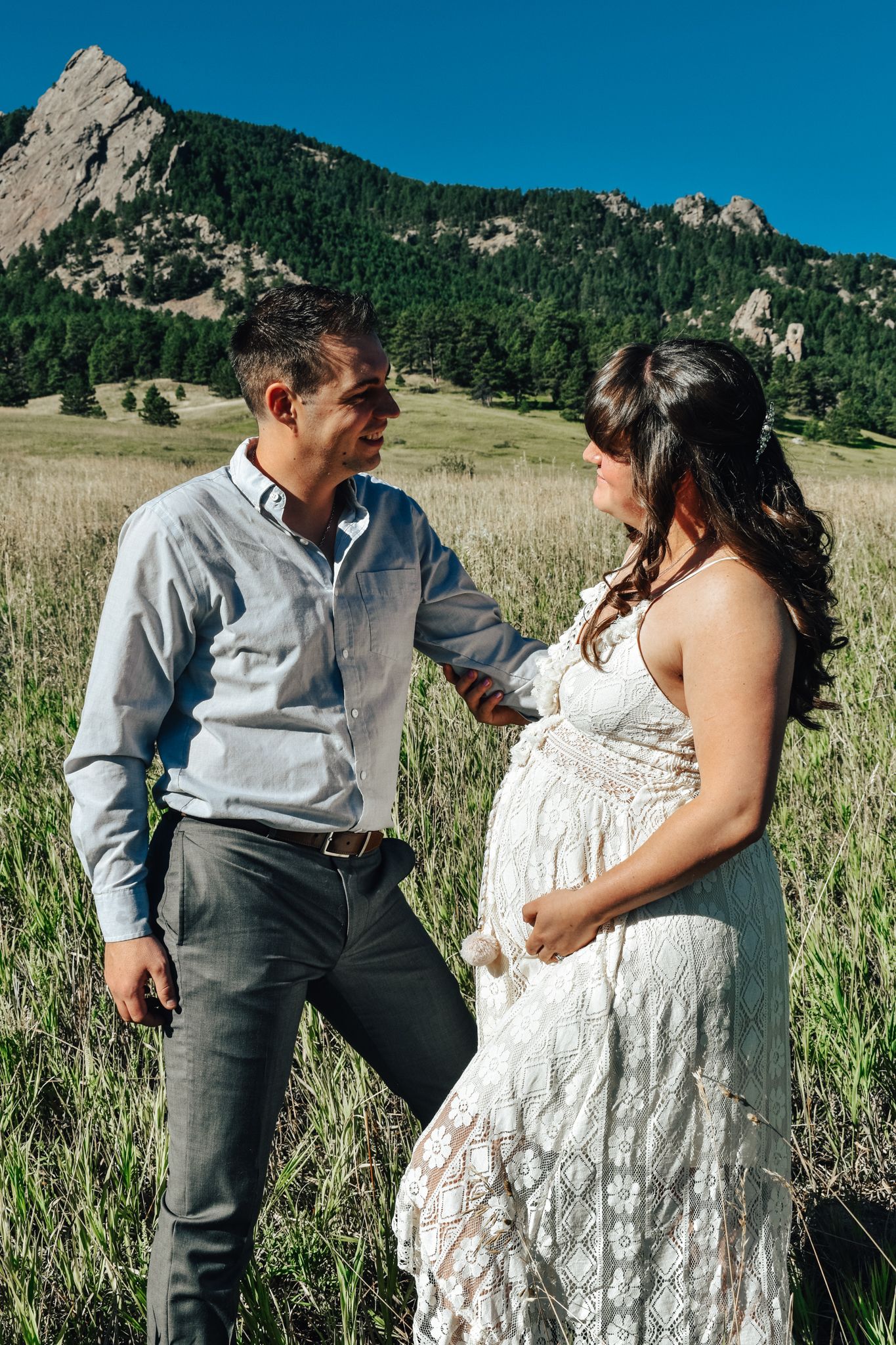 Pregnant couple standing in a green meadow with rocky mountains of Colorado on a sunny day.  Maternity Photographer in Pflugerville, Tx