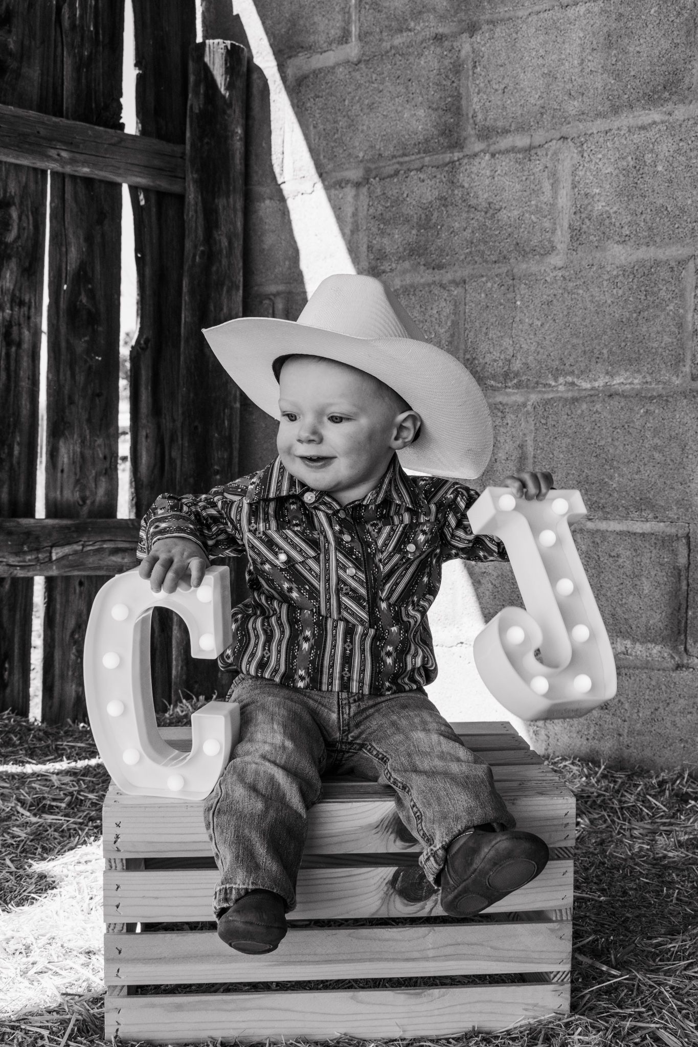 Smiling toddler in cowboy hat sits on wooden crate with marquee letters C and J. Family Photographer in Pflugerville, Tx