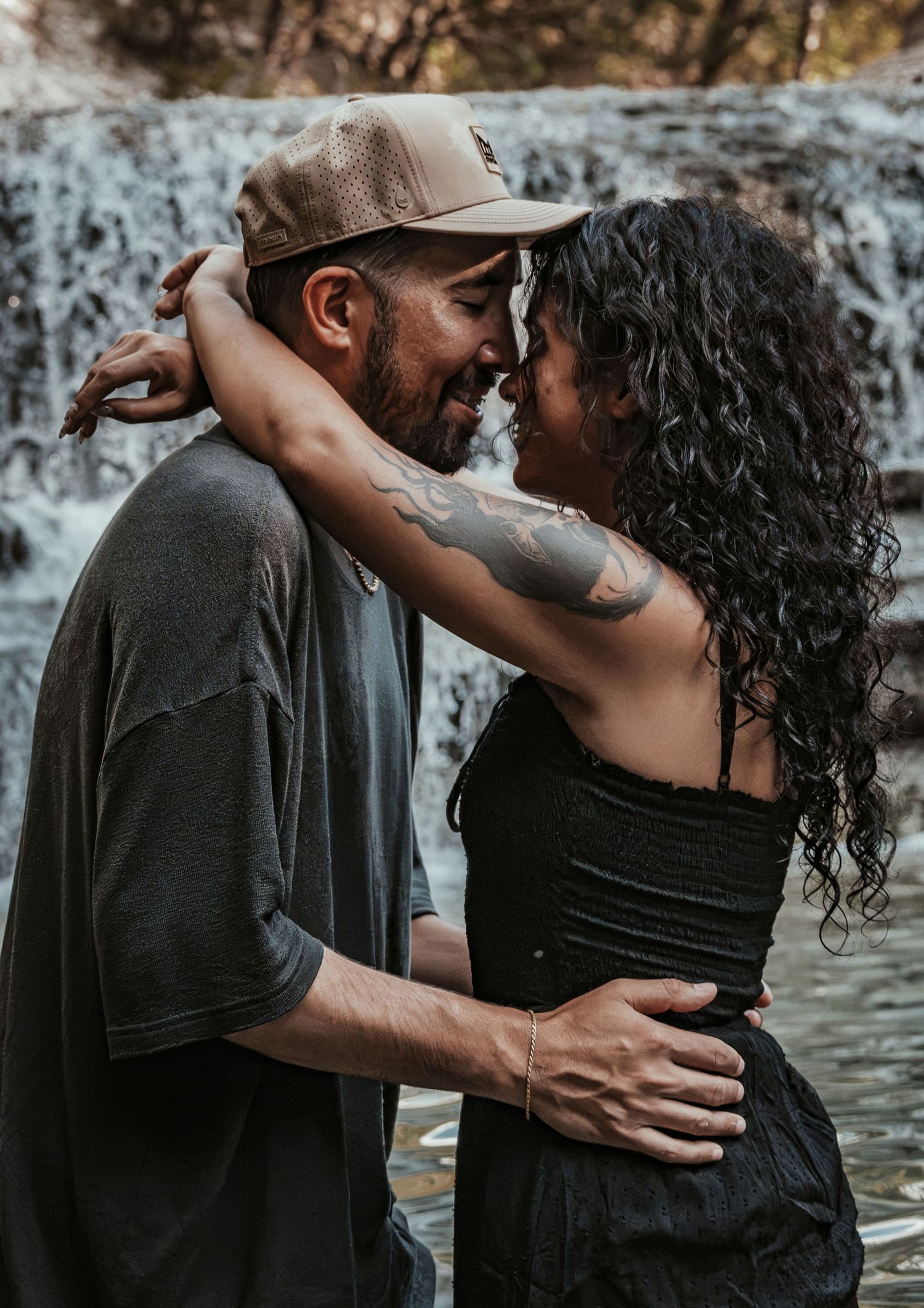 A couple embraces and almost kisses in front of a waterfall, the woman has a tattoo for a couples session.
