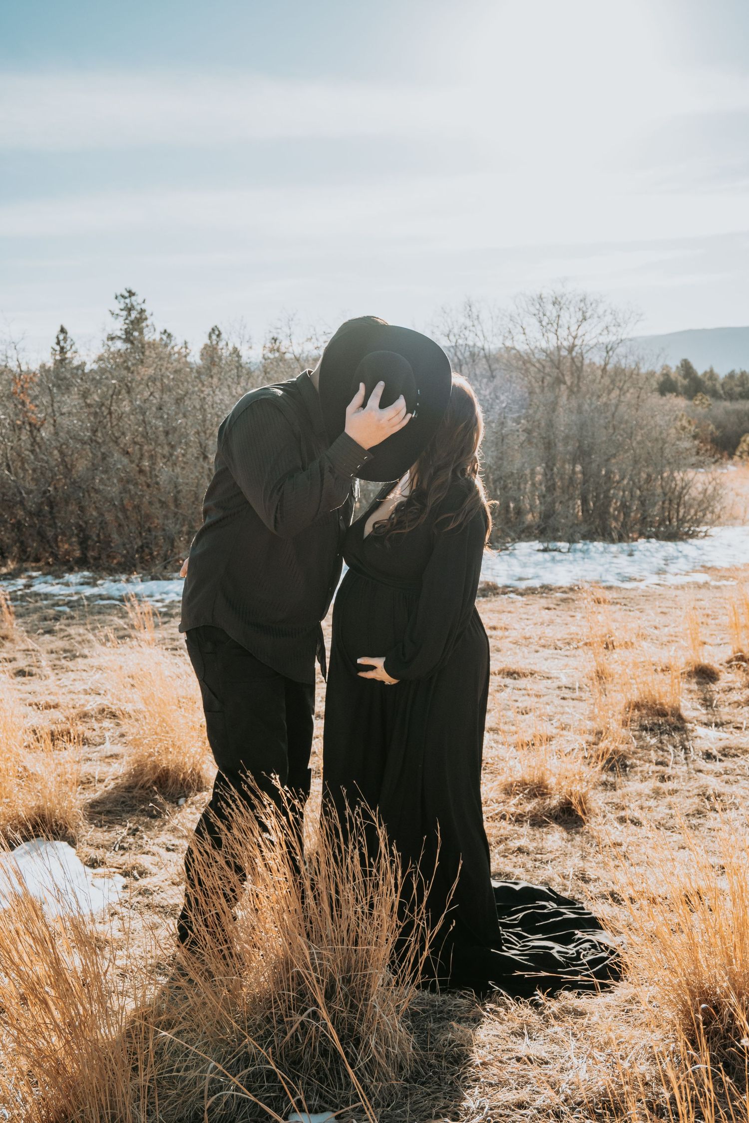 Couple kissing in a snowy winter field, both dressed in black during an outdoor maternity photoshoot.