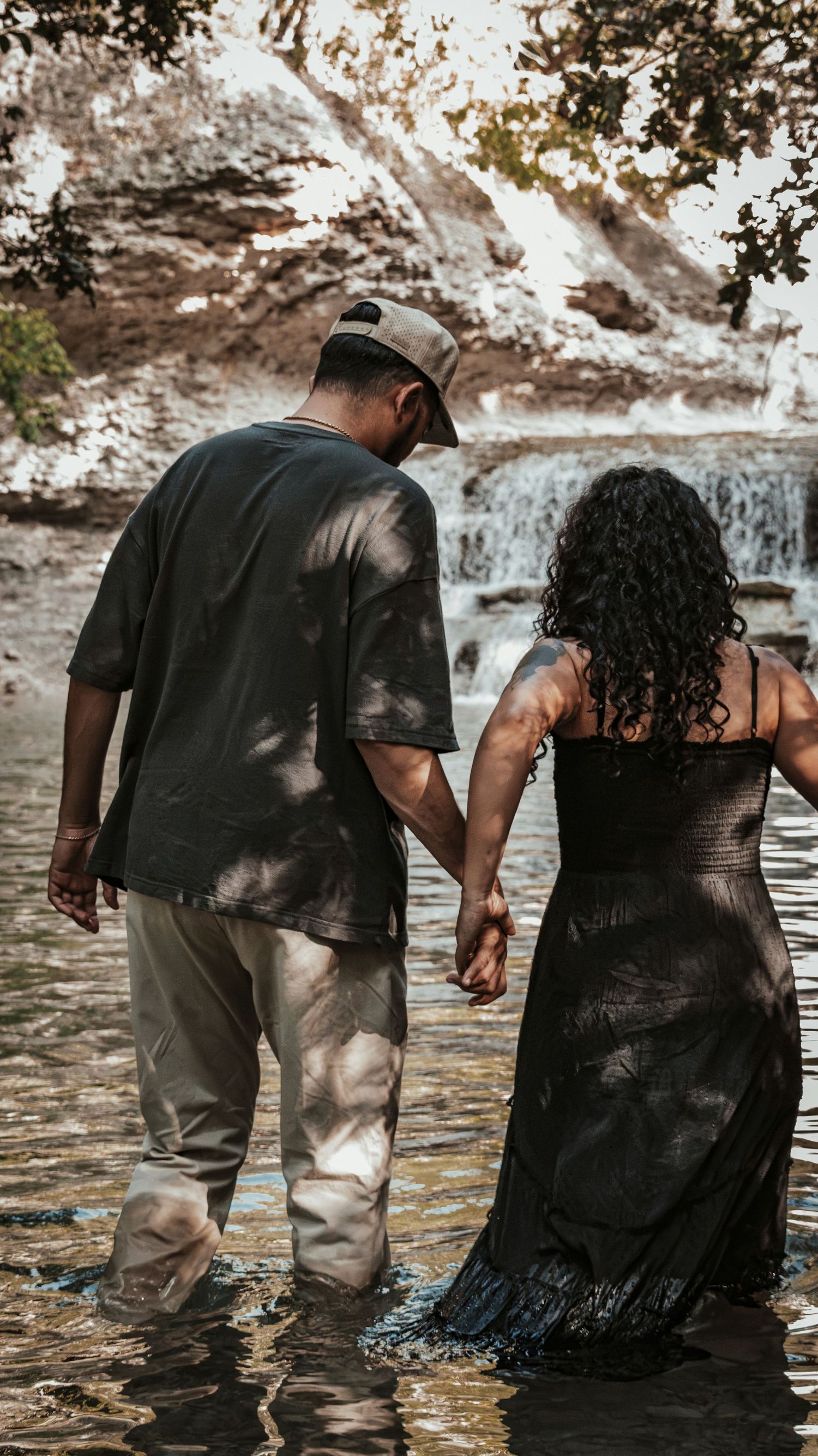 Couple holding hands wading through shallow water near a scenic waterfall in nature.