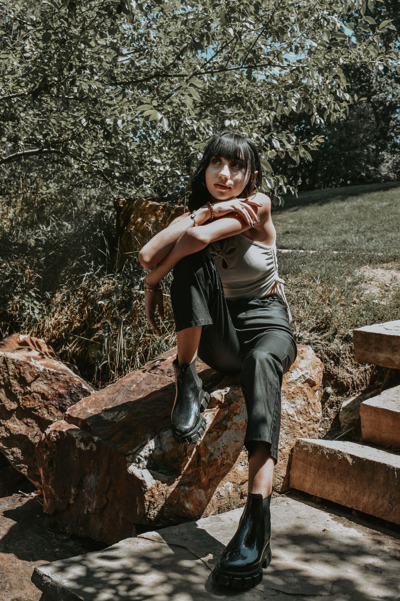 Young woman with dark bangs sits on a rock in a sunny park wearing a tank top and black pants for senior photos.