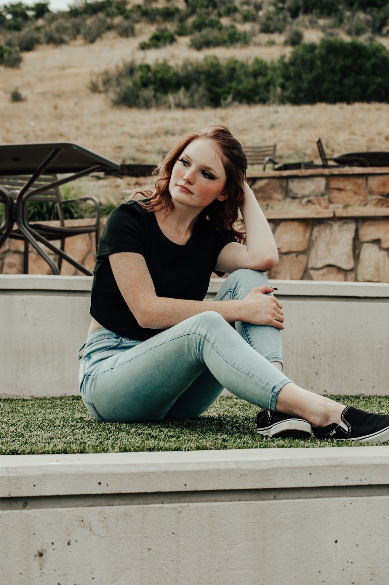 Young redhead woman in black tee and light jeans sitting on grass ledge with hand in hair outdoors for senior photos.