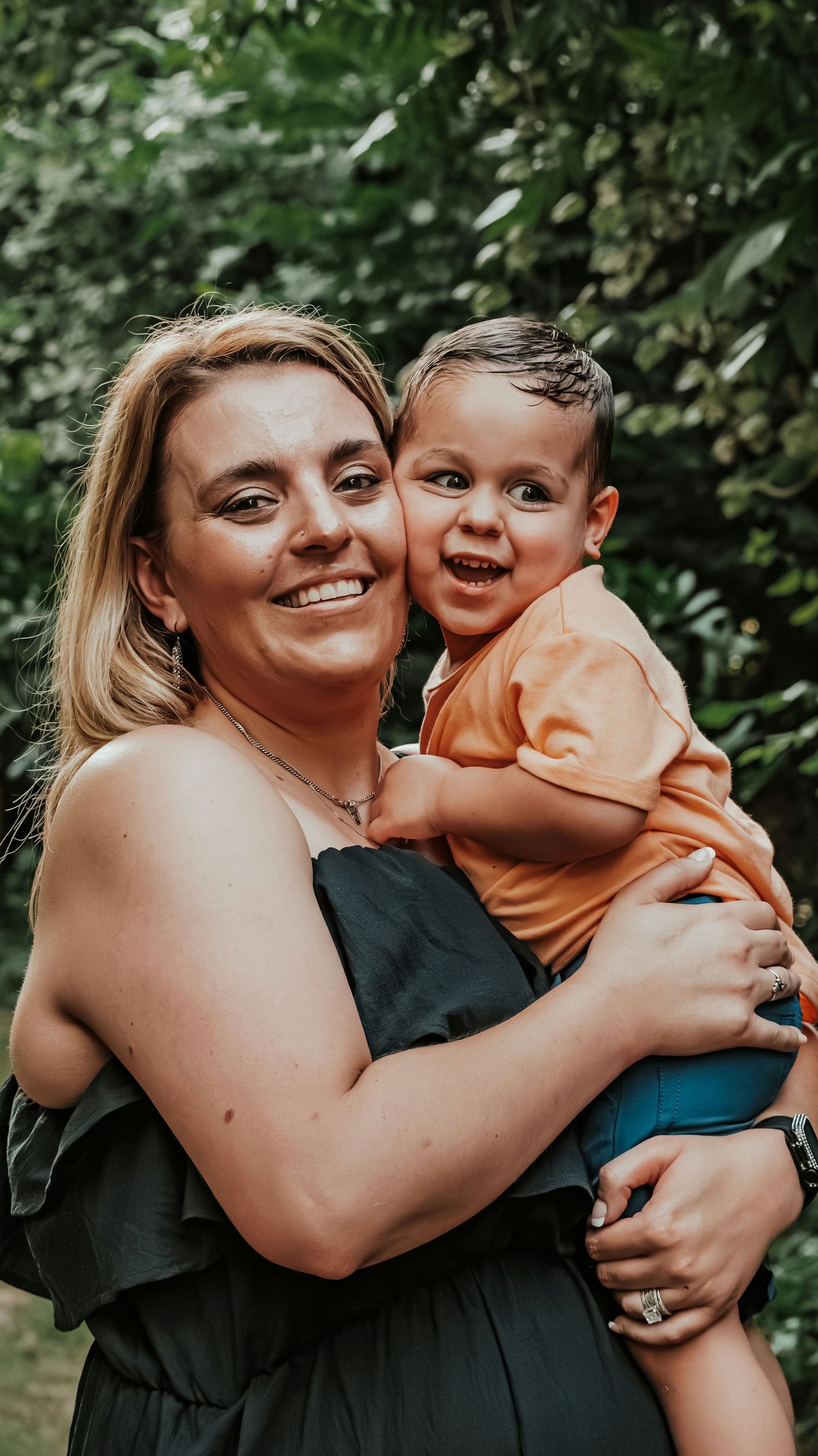 Smiling pregnant woman in black dress holding her happy toddler son in an orange shirt outdoors.