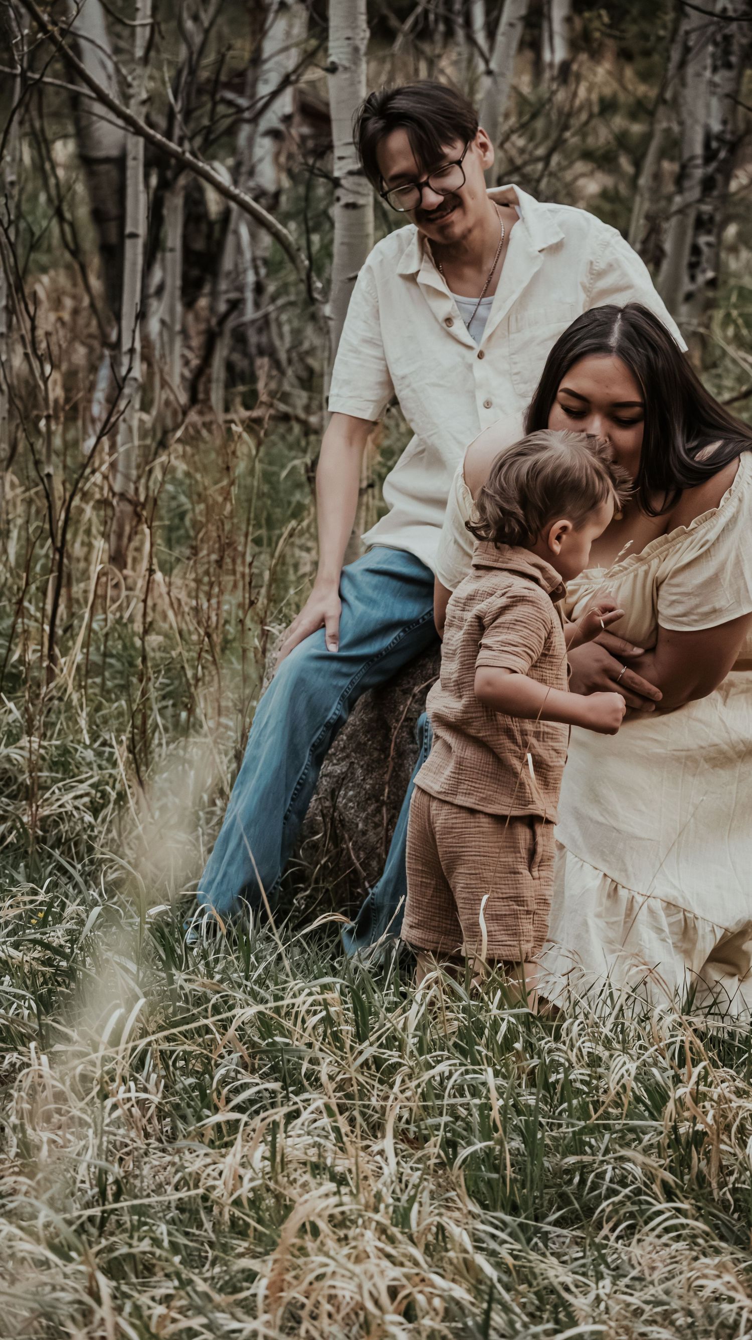 A smiling family of three shares a tender moment together in a lush grassy woodland setting.
