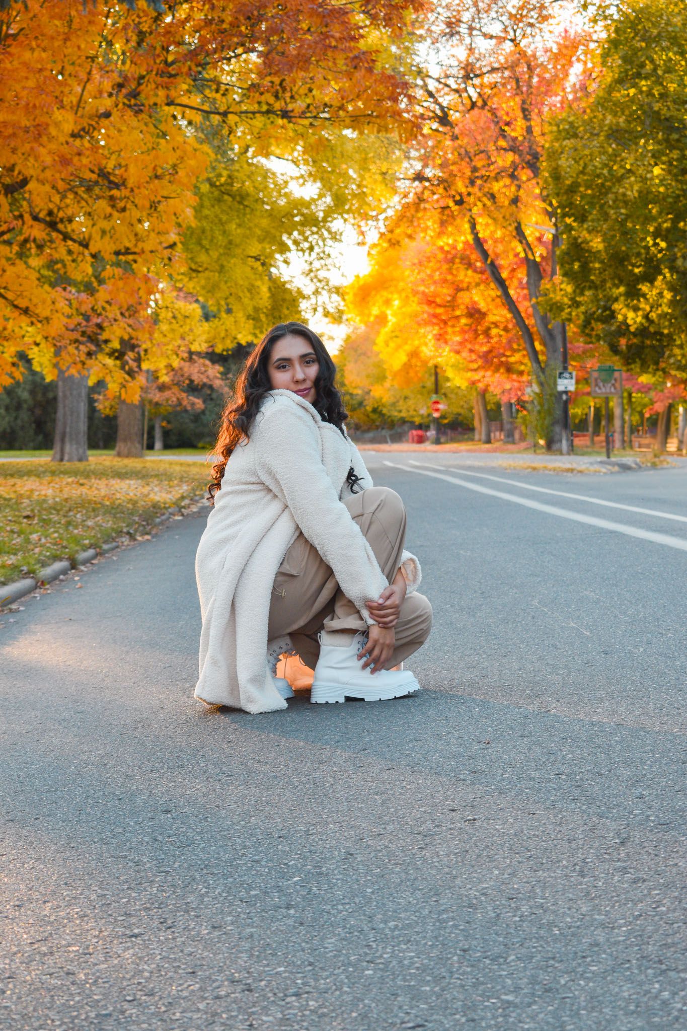 Woman in cream coat crouching on road with stunning orange and red autumn trees behind her for senior photos.