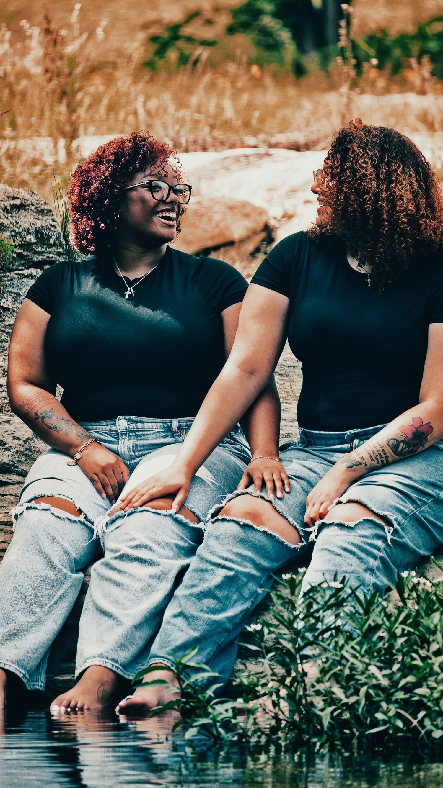 Two curly-haired women in black tops and ripped jeans laugh together sitting by water for senior photos.