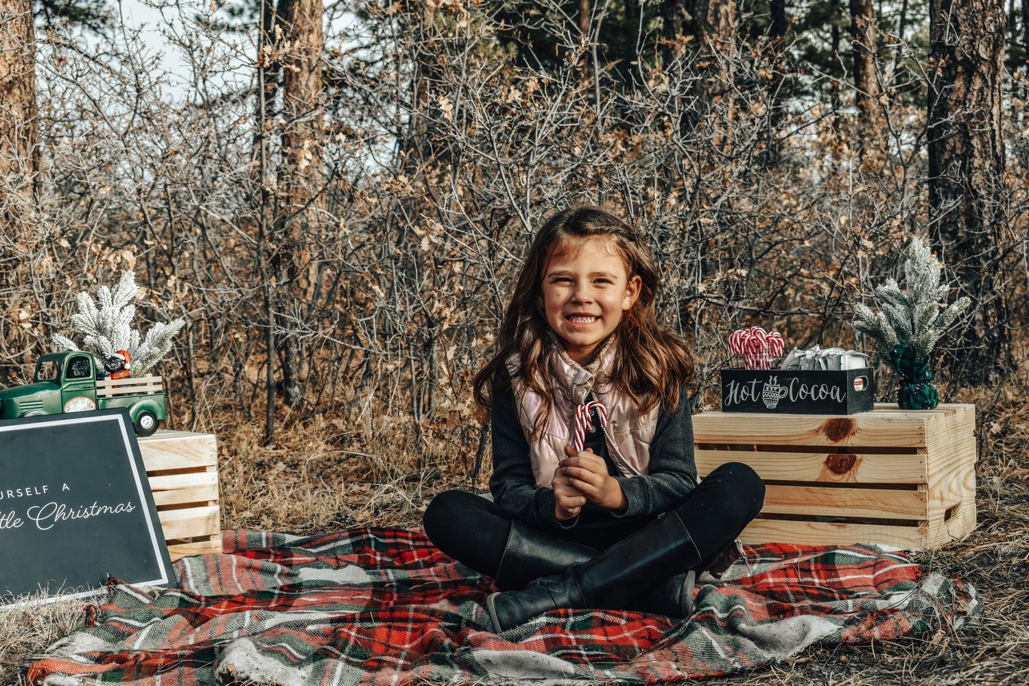 A smiling girl with long brown hair sits cross-legged on a plaid blanket with Christmas props in a forest.