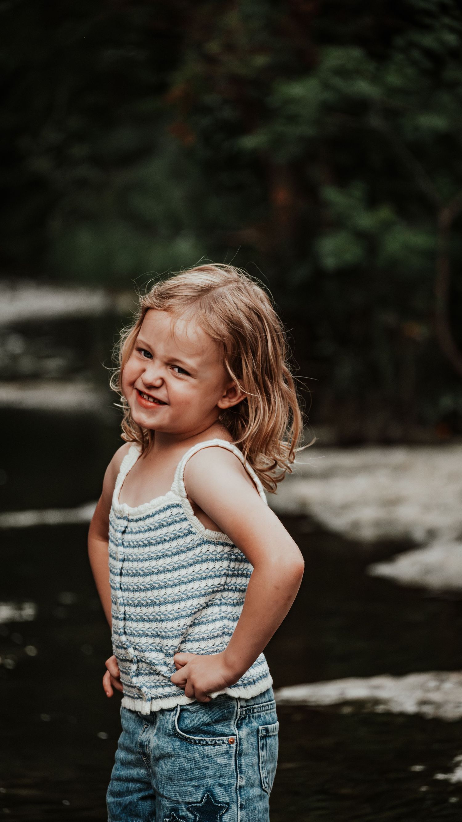 A confident little girl in a striped tank top and jeans poses with hands on hips by a river.