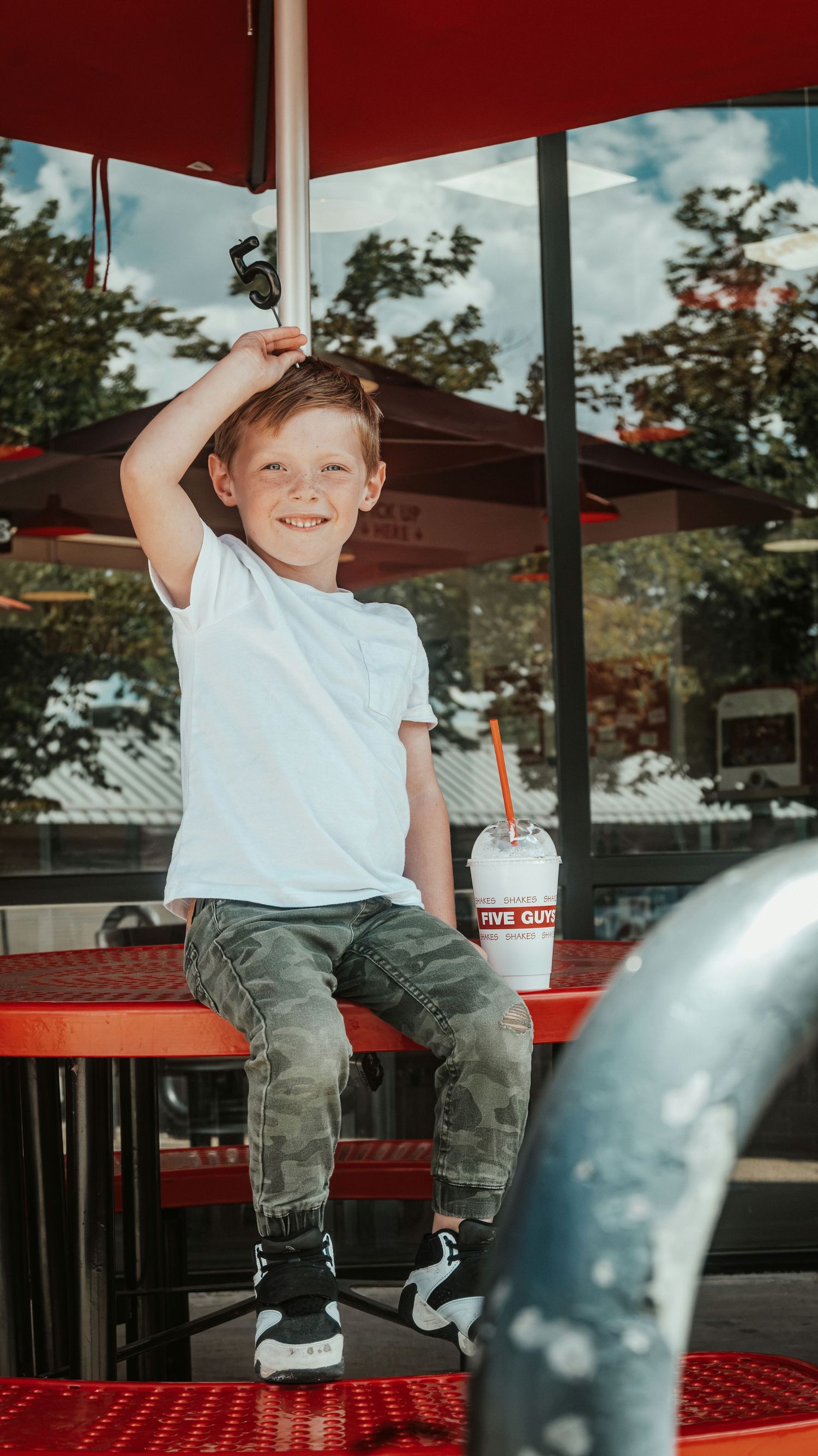 Smiling boy sitting on red outdoor table at Five Guys holding a milkshake.