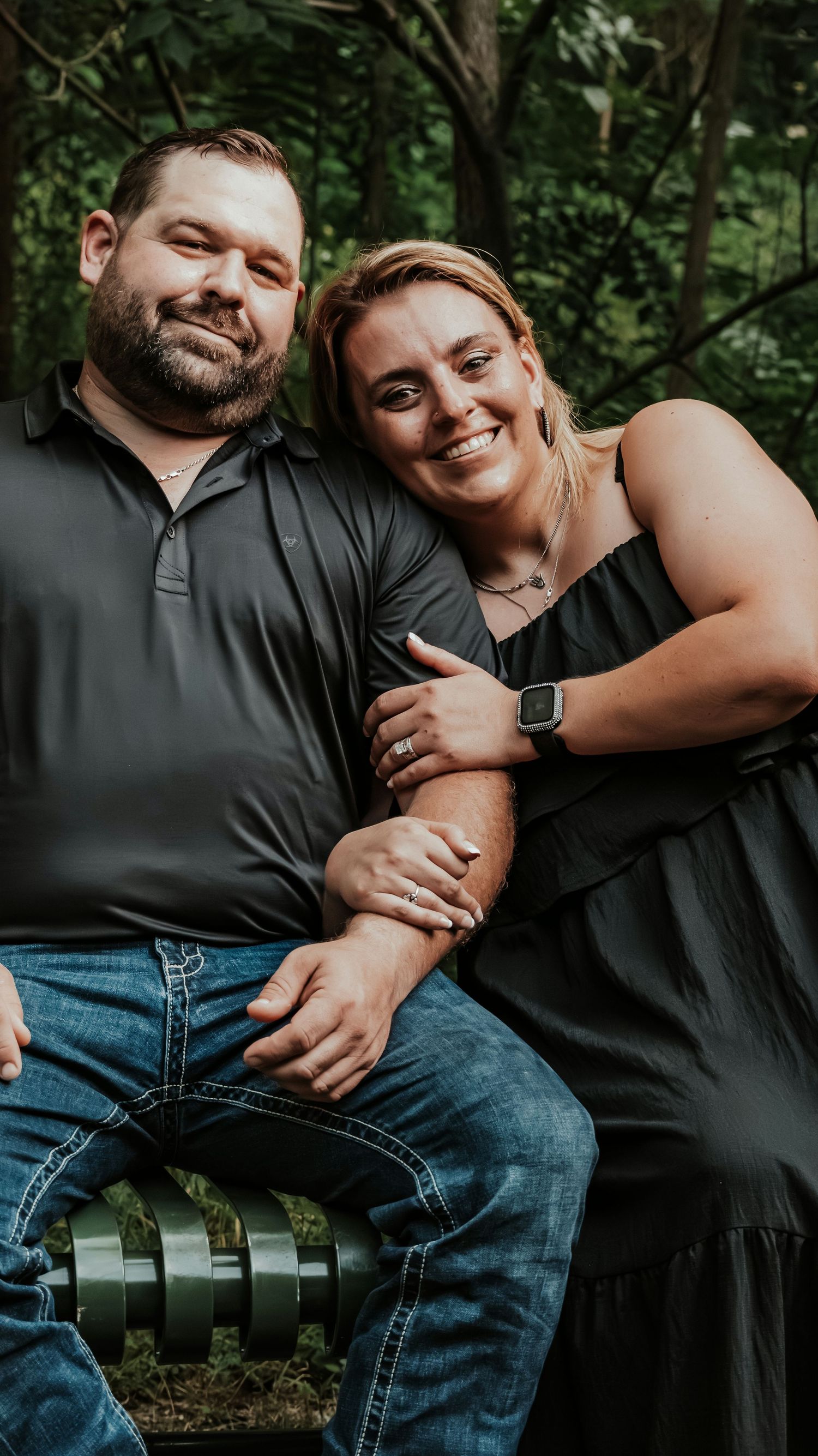 Smiling couple sitting together outdoors, woman in black dress leaning on man in black polo and jeans.