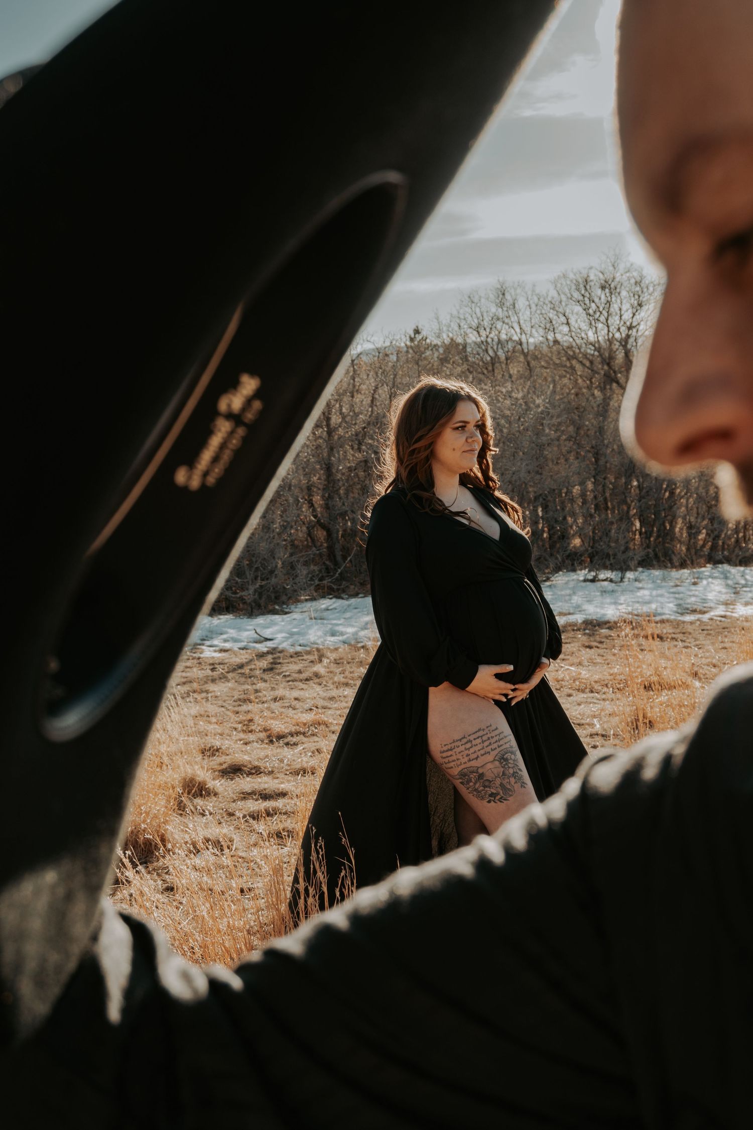 Pregnant woman in black coat cradles baby bump in golden field while partner stands in foreground.