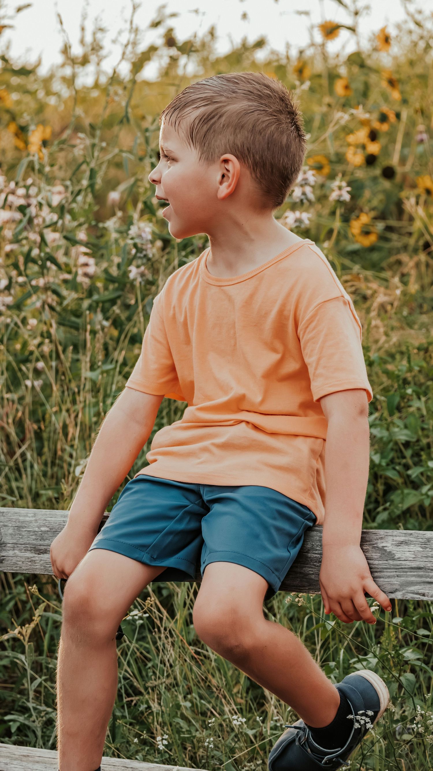 Young boy in orange shirt and blue shorts sits on wooden fence surrounded by wildflowers in golden light.