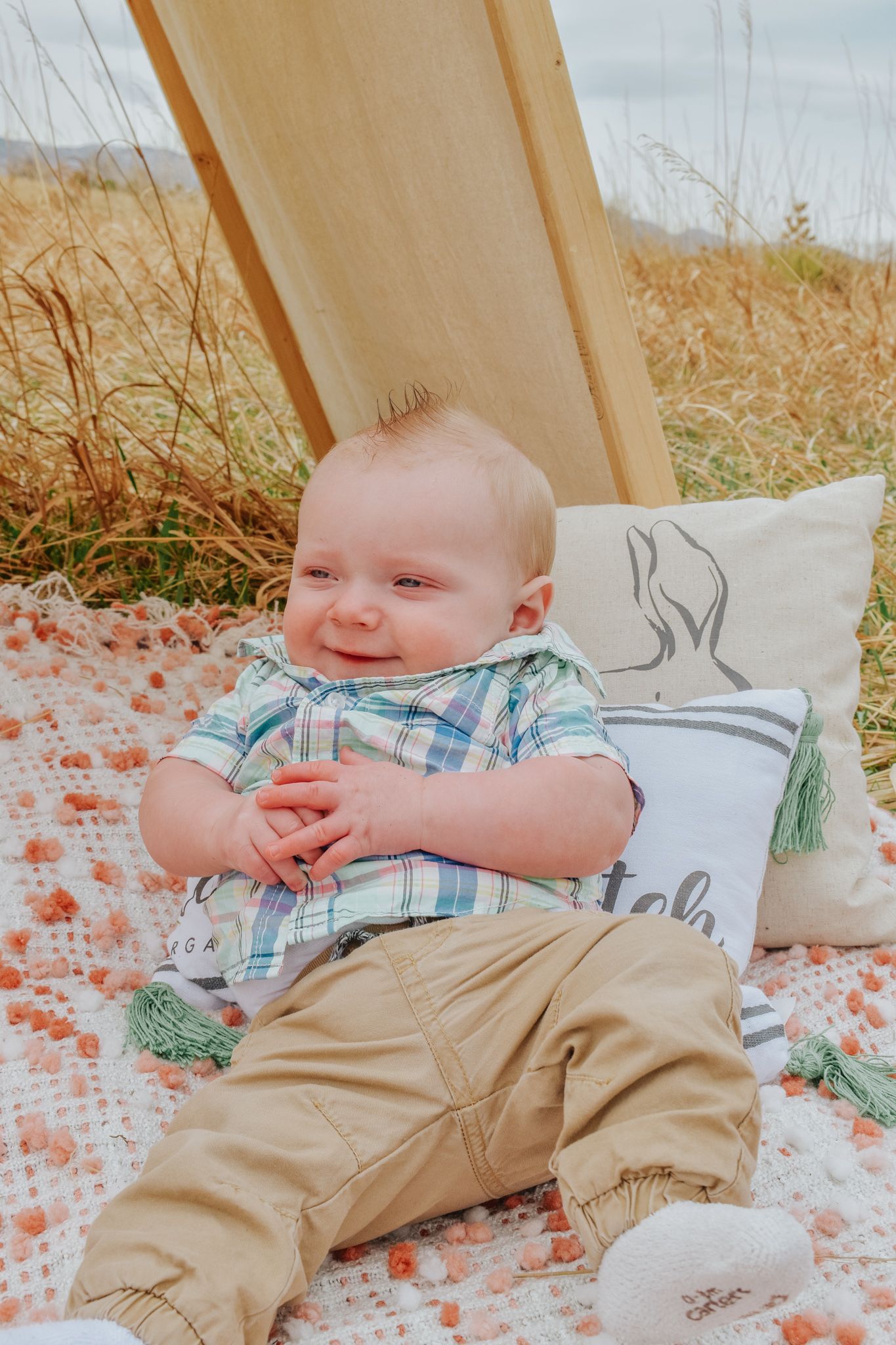 Smiling baby boy in plaid shirt and khaki pants sitting on a blanket outdoors near tall grass. Family Photographer in Pflugerville, Tx