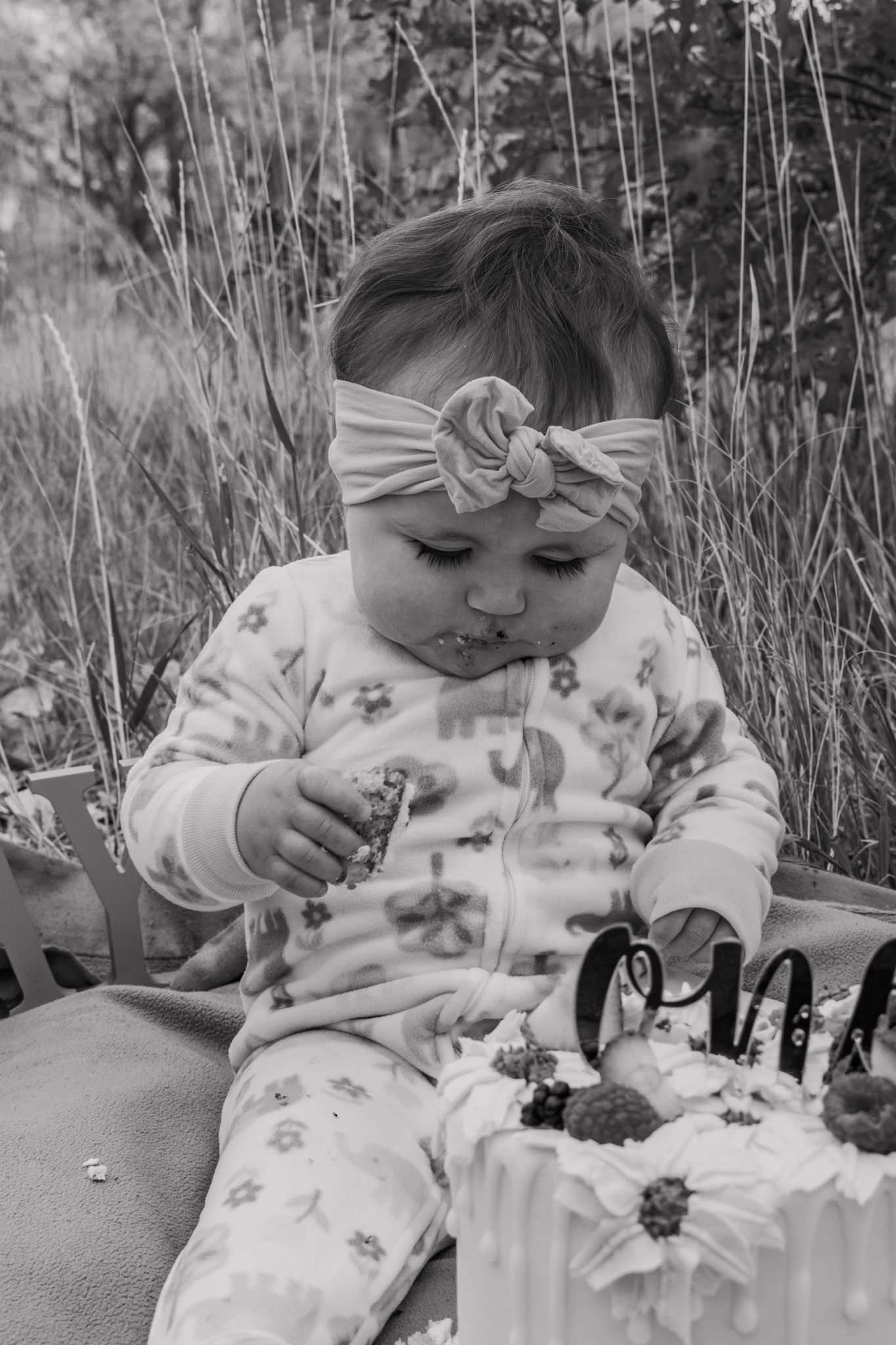 Black and white photo of a baby girl with bow headband exploring her first birthday smash cake. Family Photographer in Pflugerville, Tx