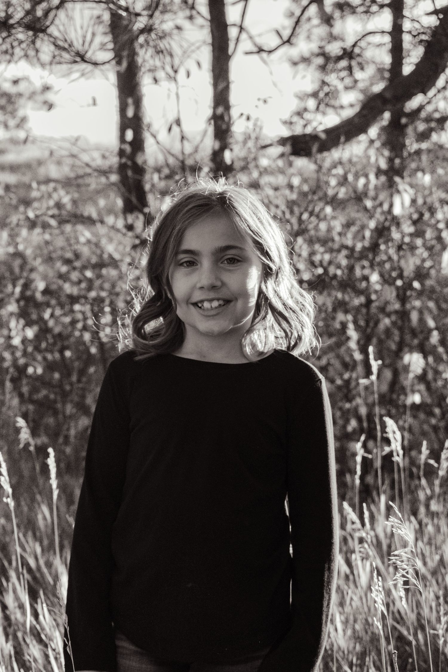 Black and white portrait of a smiling young girl with wavy hair in a black top outdoors. Family Photographer in Pflugerville, Tx