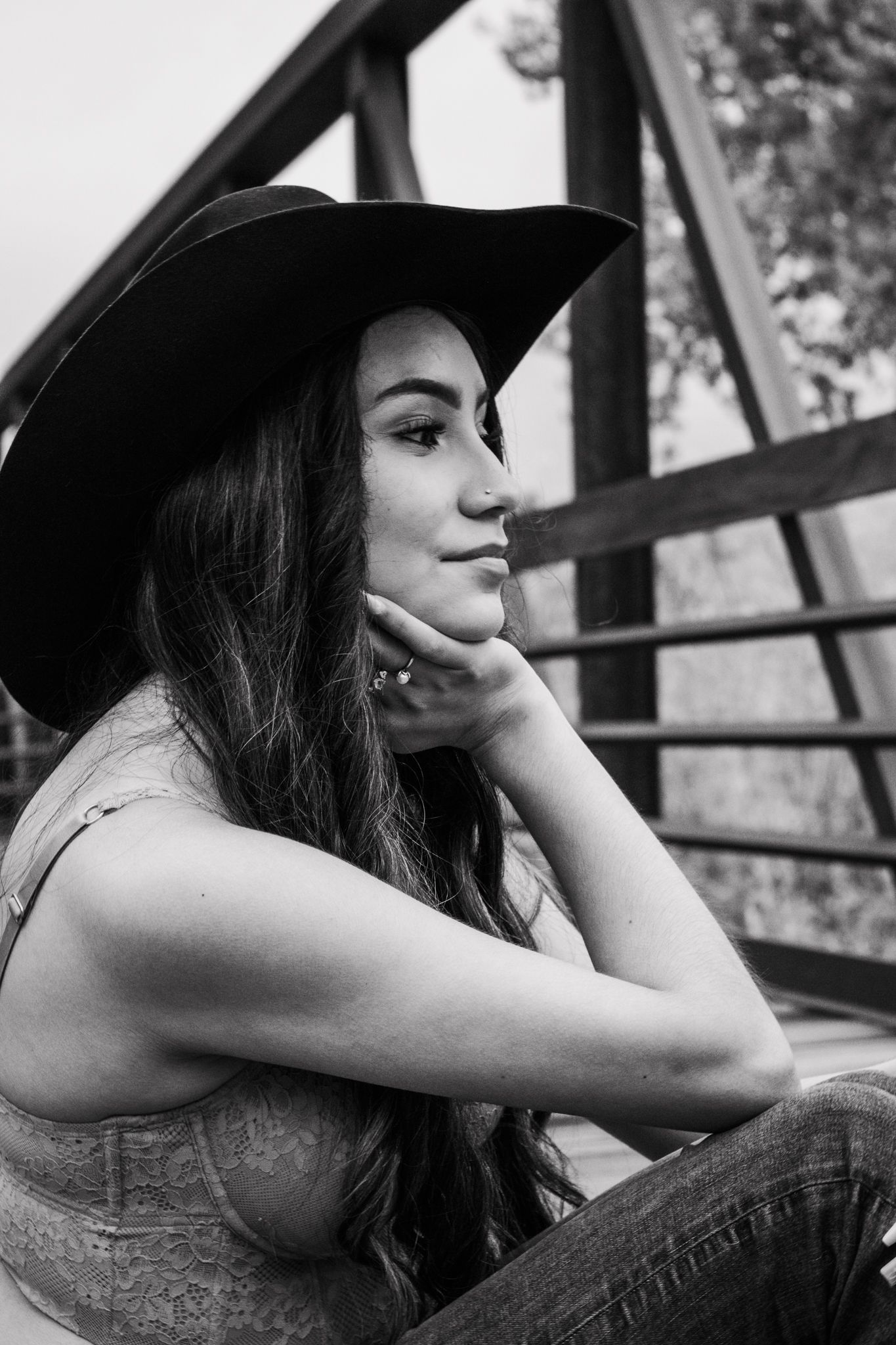 Black and white portrait of young woman in wide-brim hat sitting on a bridge, resting chin on hand for senior photos.