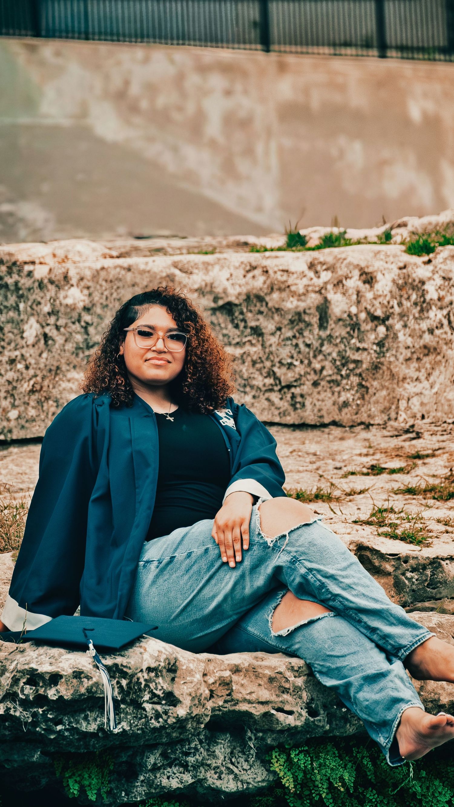 Young woman in graduation gown with glasses sitting relaxed on a stone ledge outdoors for senior photos.