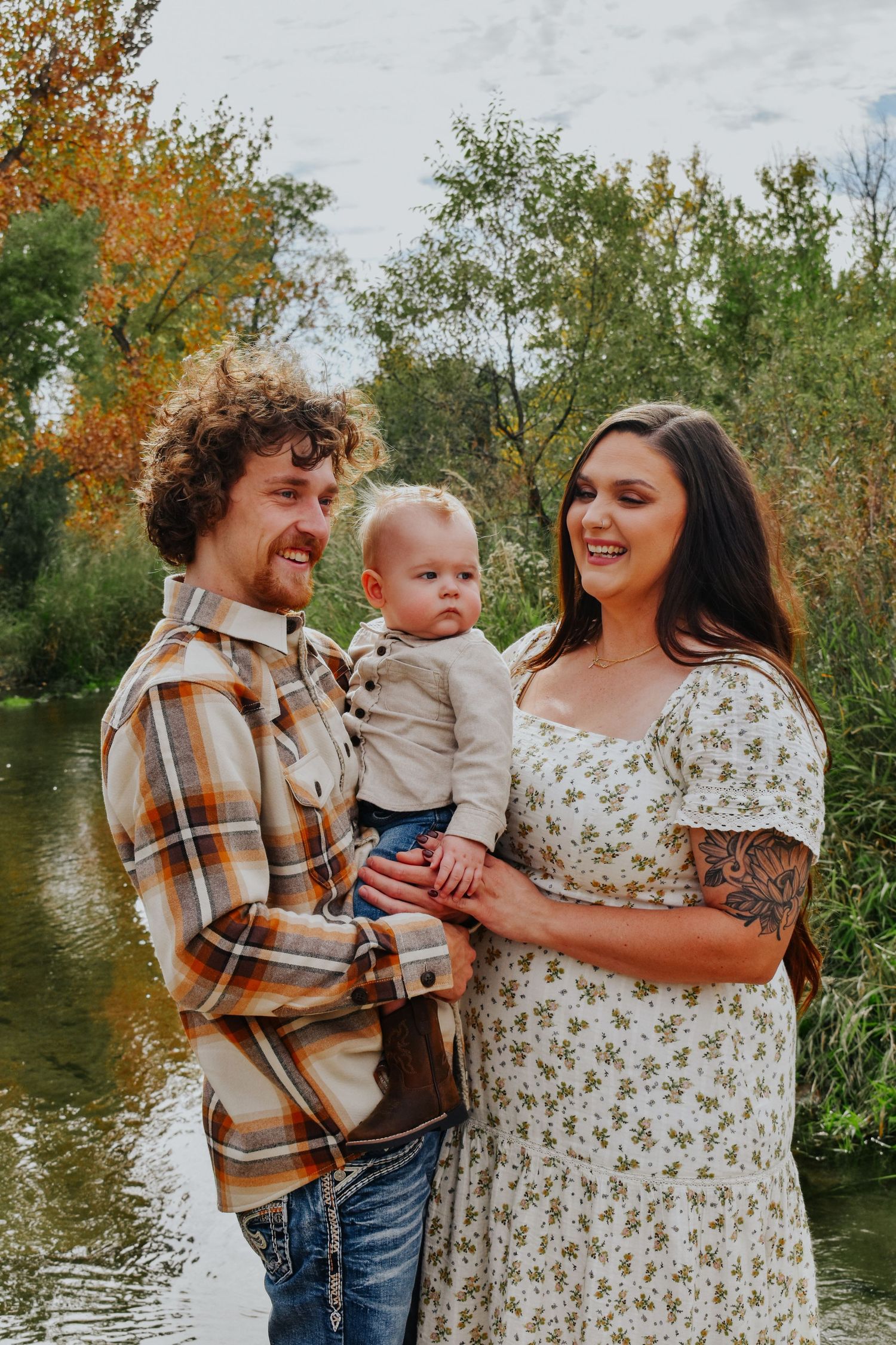 Happy young family with father in plaid shirt and mother in floral dress holding baby by river.