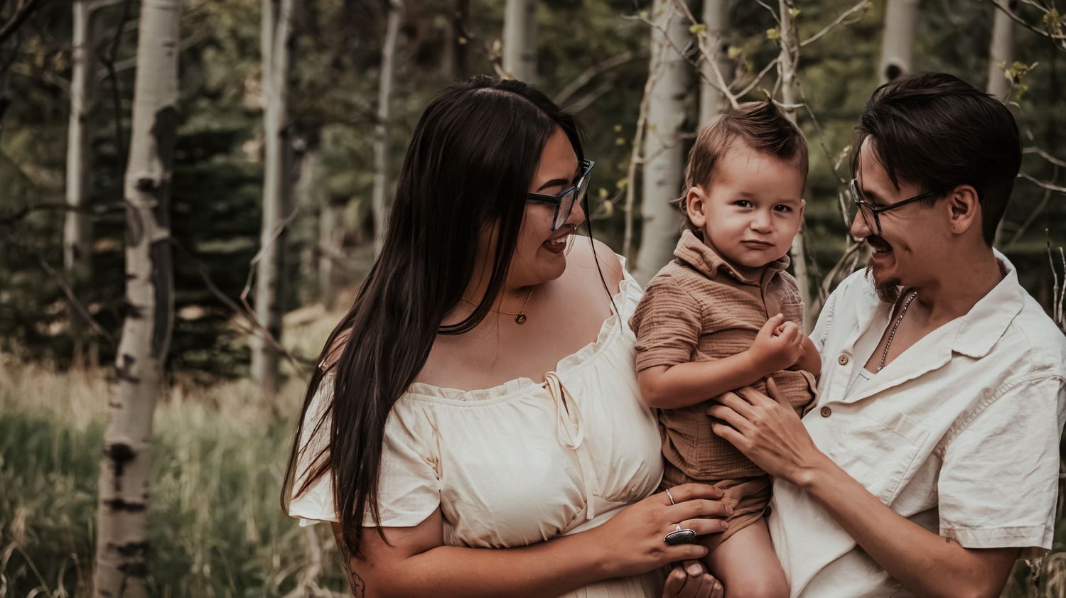 Family portrait with parents holding baby boy among birch trees in a natural outdoor setting.
