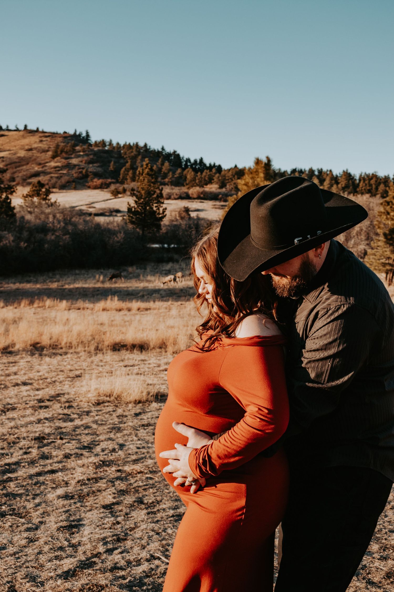 Couple shares tender moment, man in cowboy hat embraces pregnant woman in red dress.