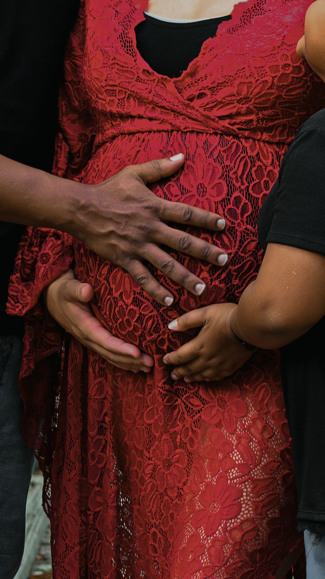 Close-up of two pairs of hands cradling pregnant belly in red lace maternity dress.