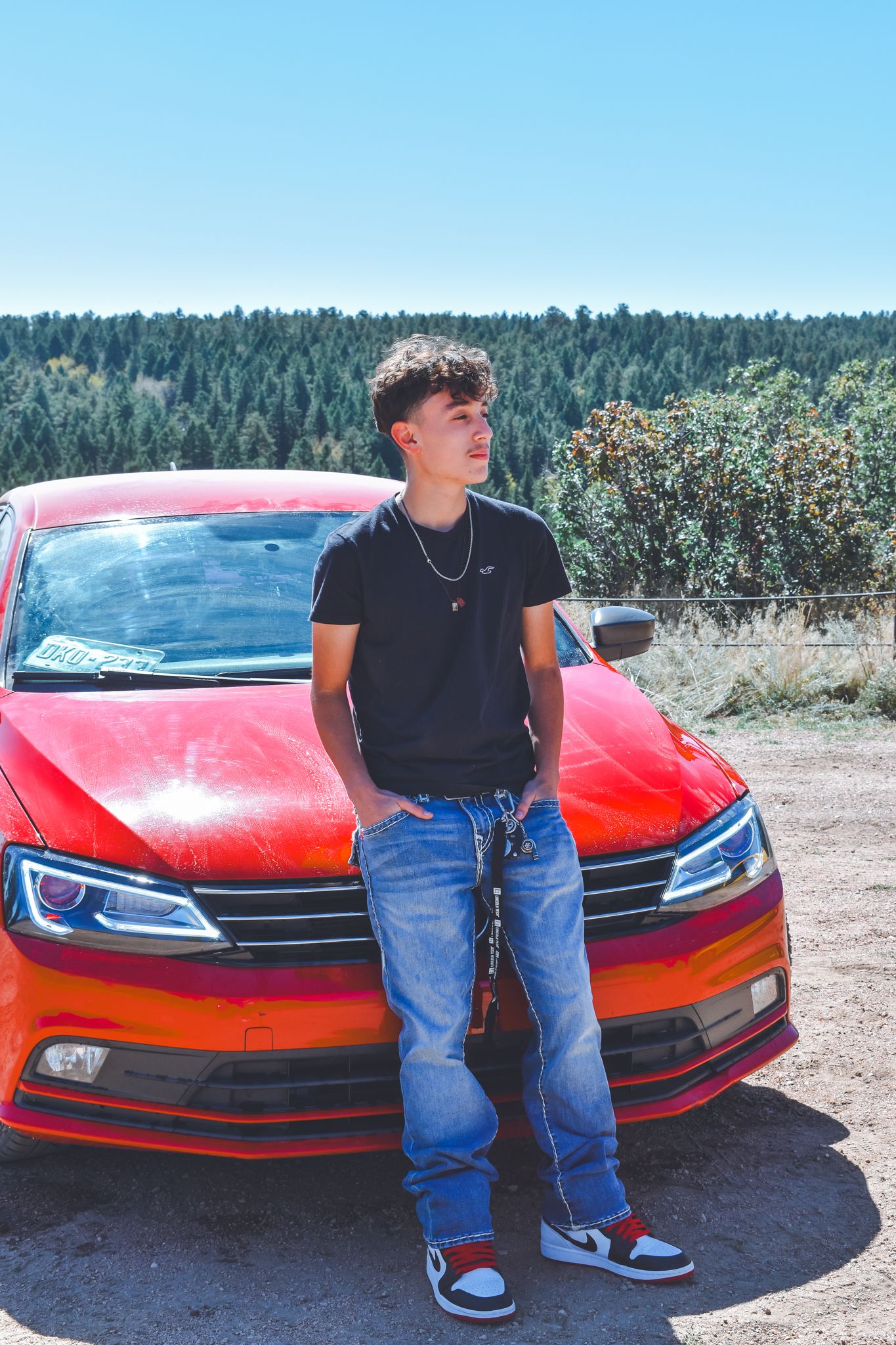 Young man in black tee and jeans leaning on a red Volkswagen car with forest backdrop for senior photos.