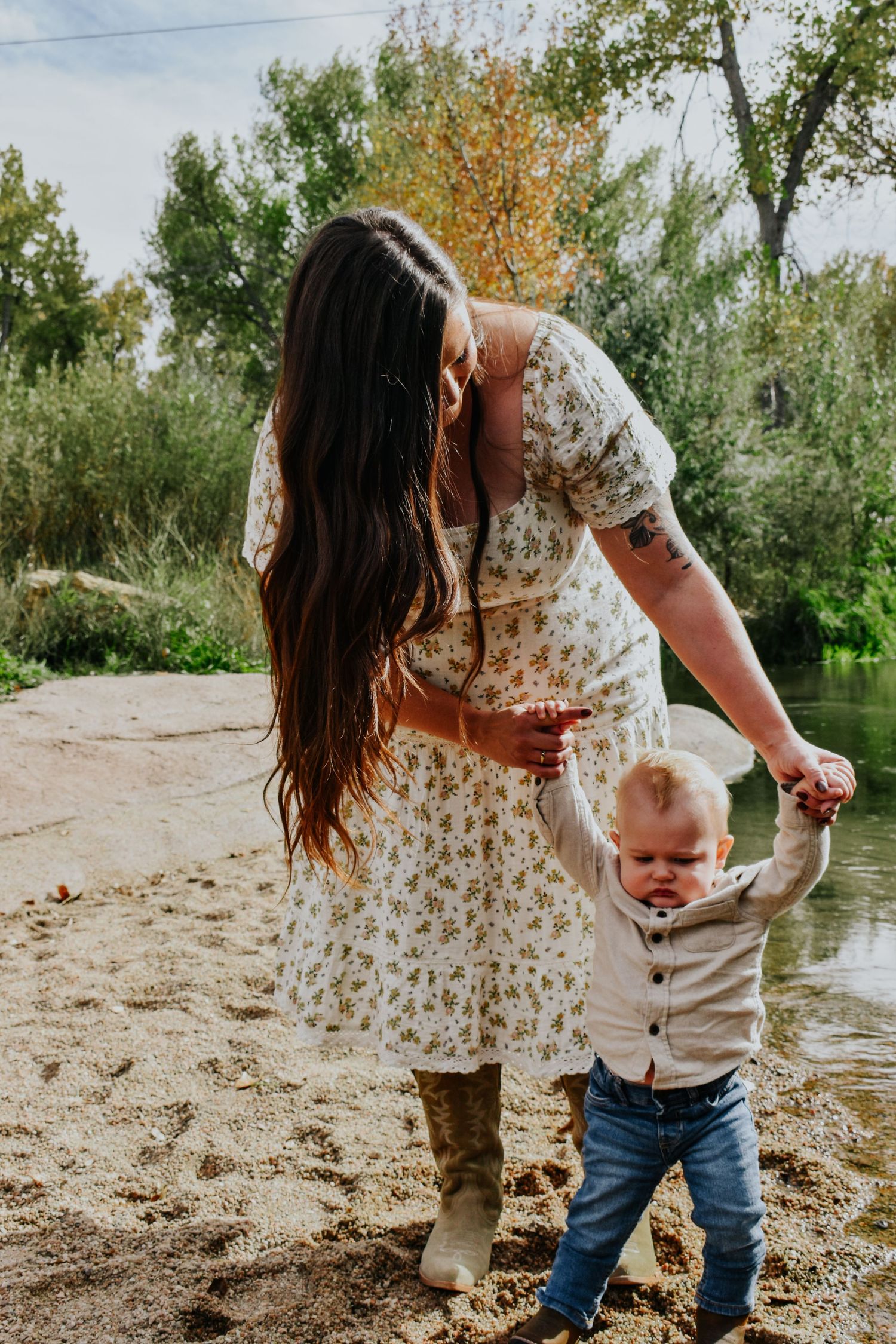 Mother in floral dress helping toddler walk along sandy riverbank outdoors.