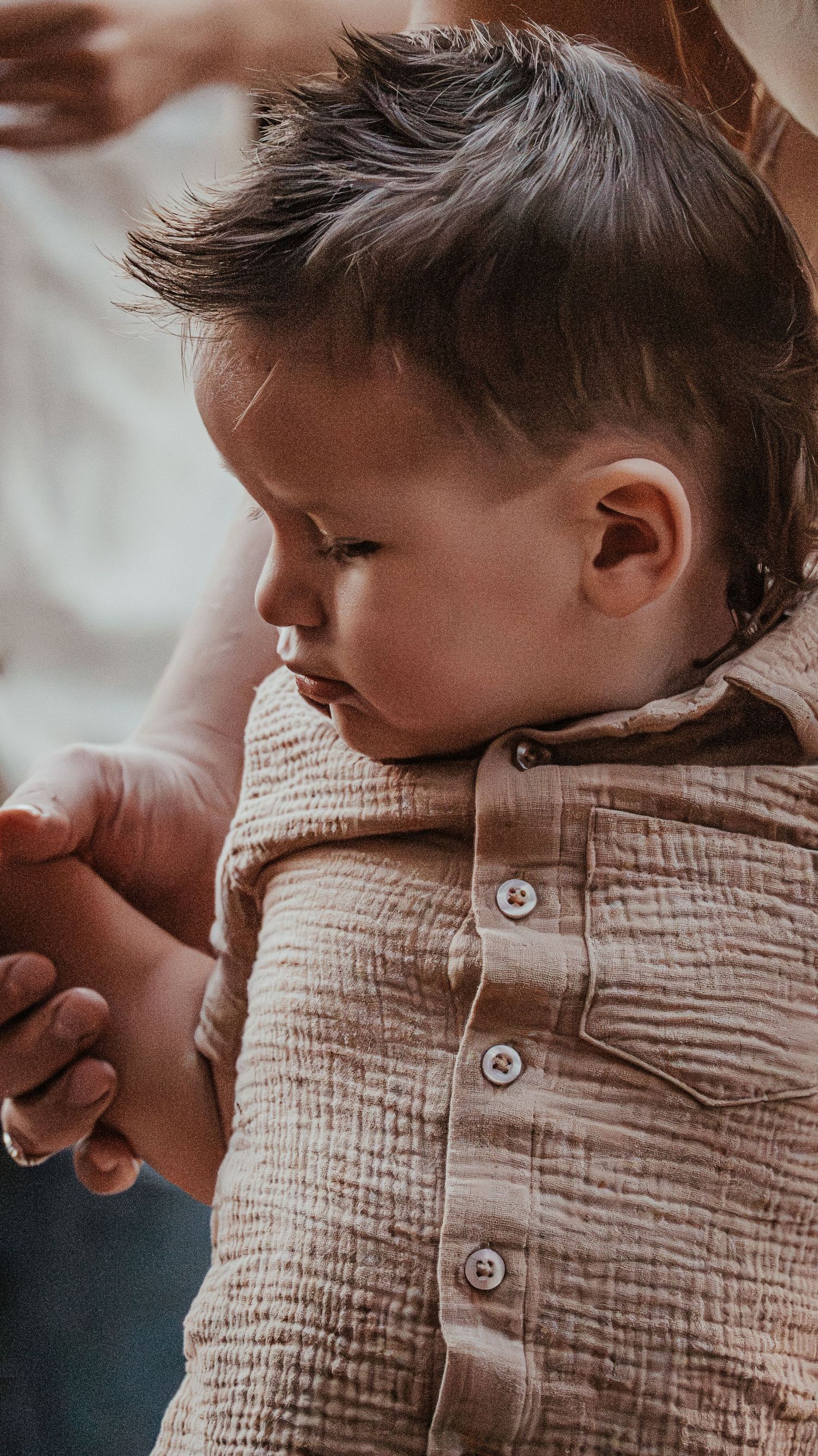 Close-up of baby boy in brown linen shirt being gently held by parent's caring hands.