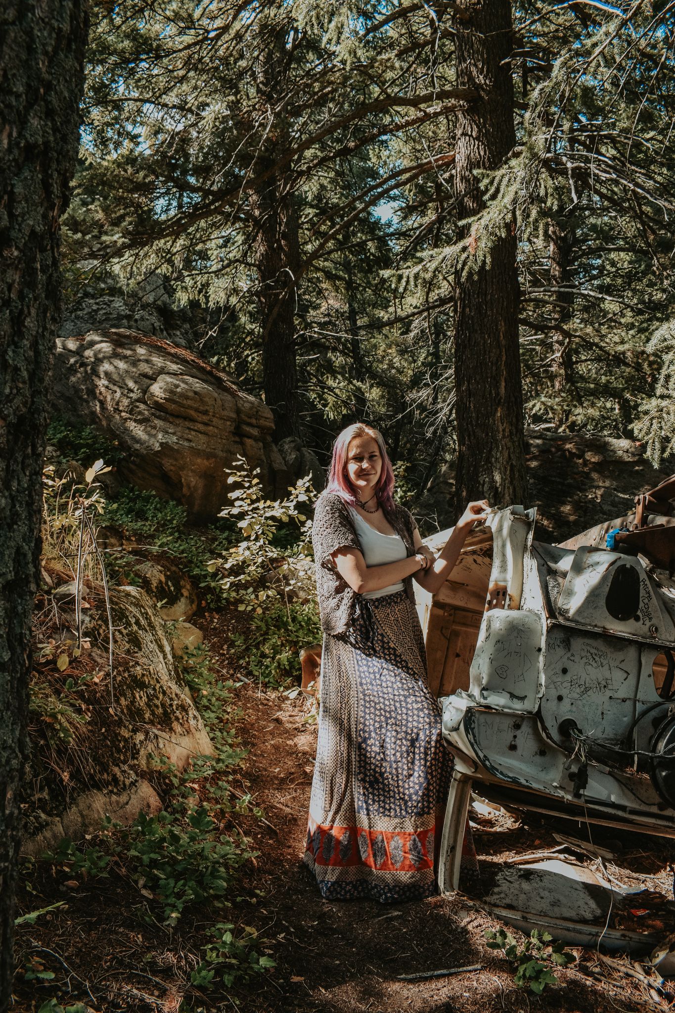Woman in flowing boho skirt stands among pine trees in sunlit forest setting for senior photos.