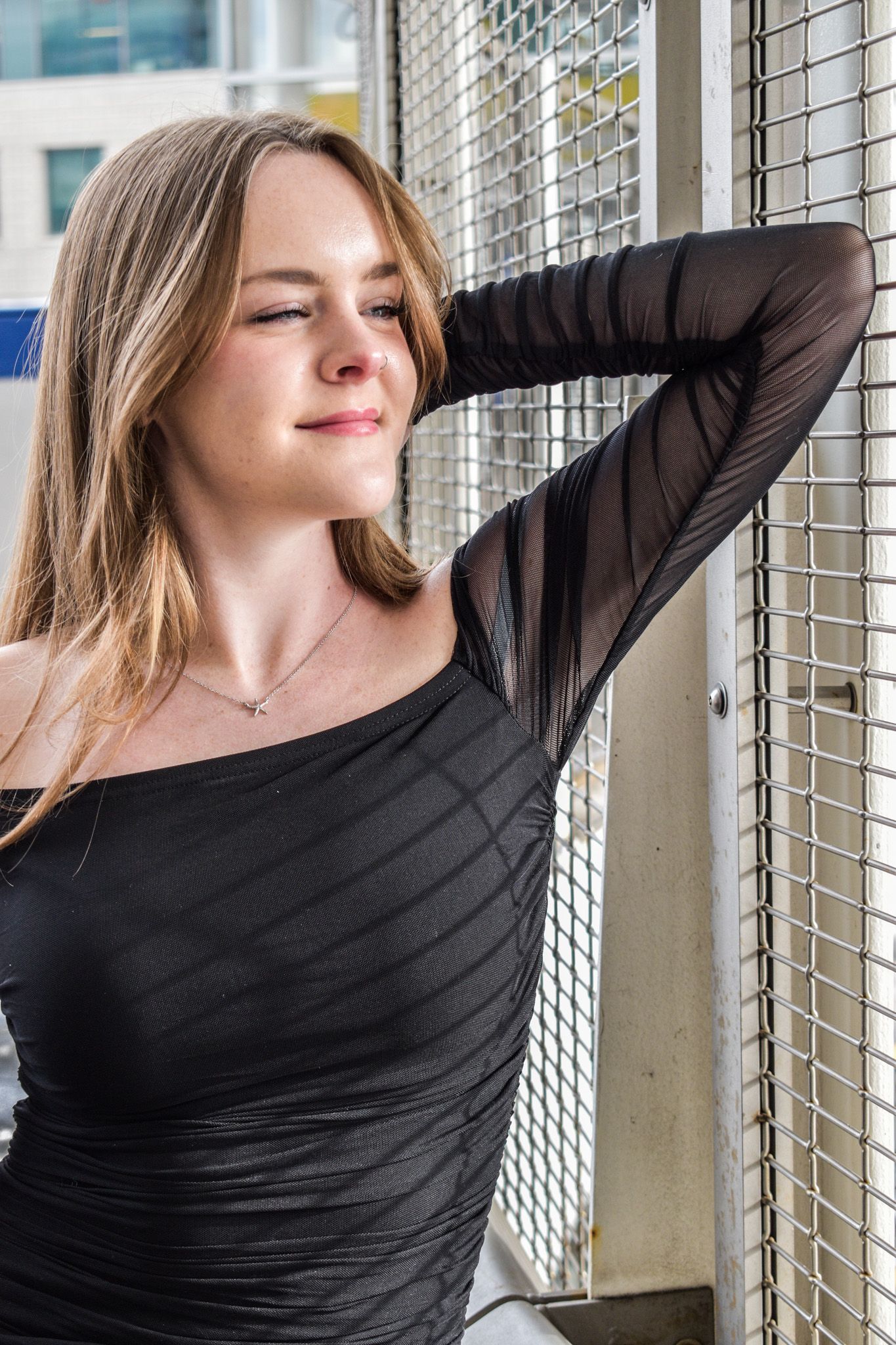 Young woman in black off-shoulder dress leaning against metal mesh fence with soft smile outdoors for senior photos.