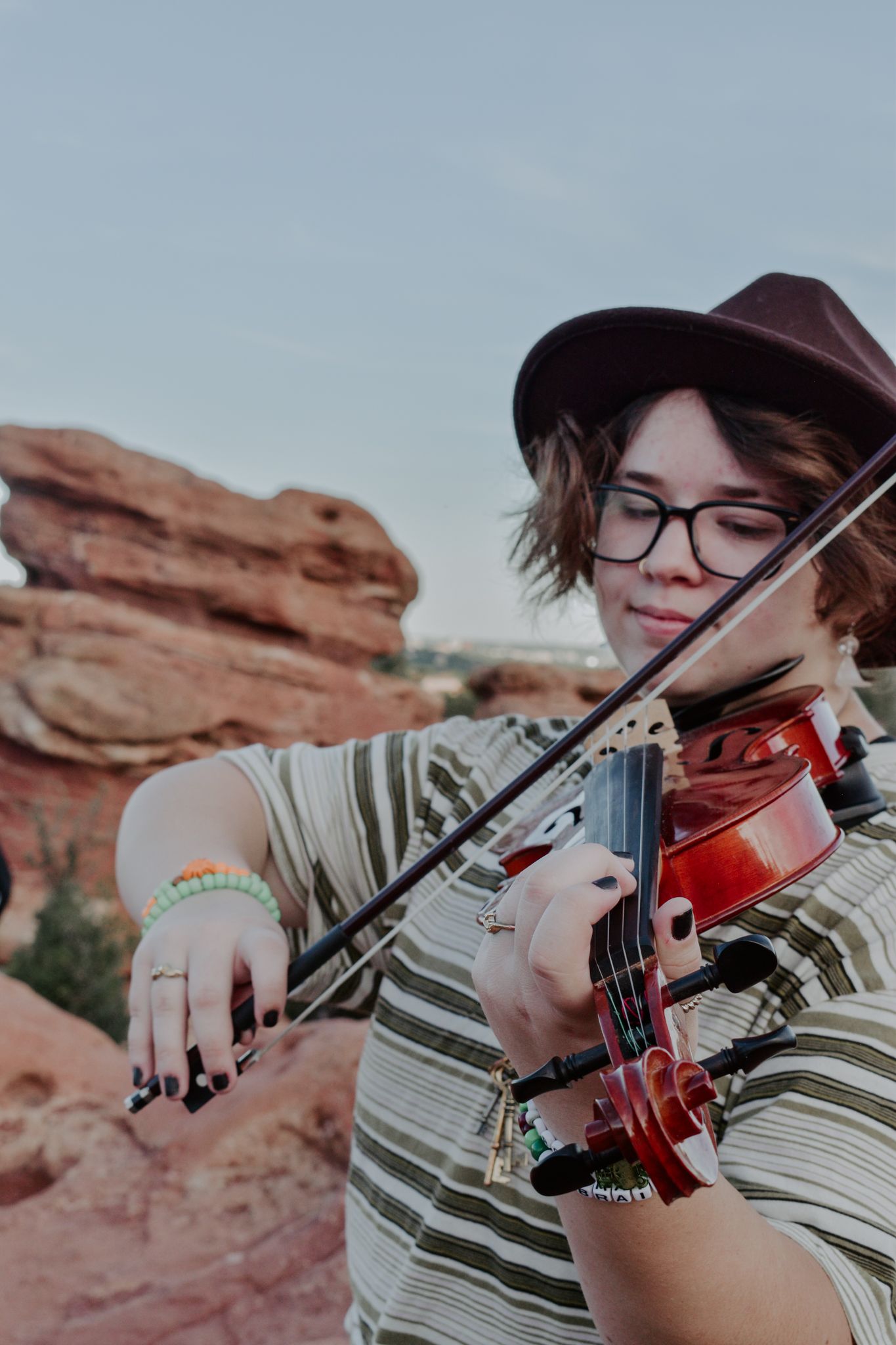 Person playing violin outdoors near red rock formations, wearing a striped shirt, hat, and glasses for senior photos.
