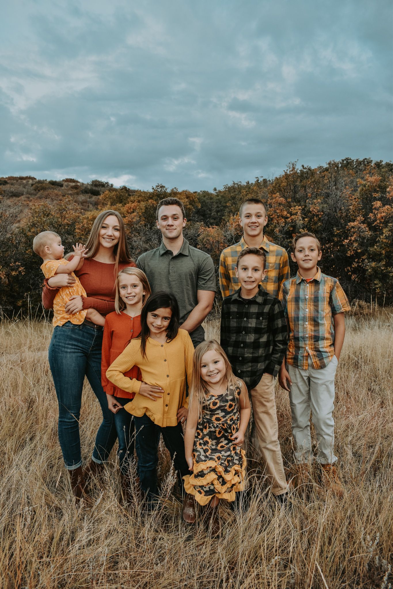 Group portrait of eight children in fall colors posing in a golden grass field with autumn trees.