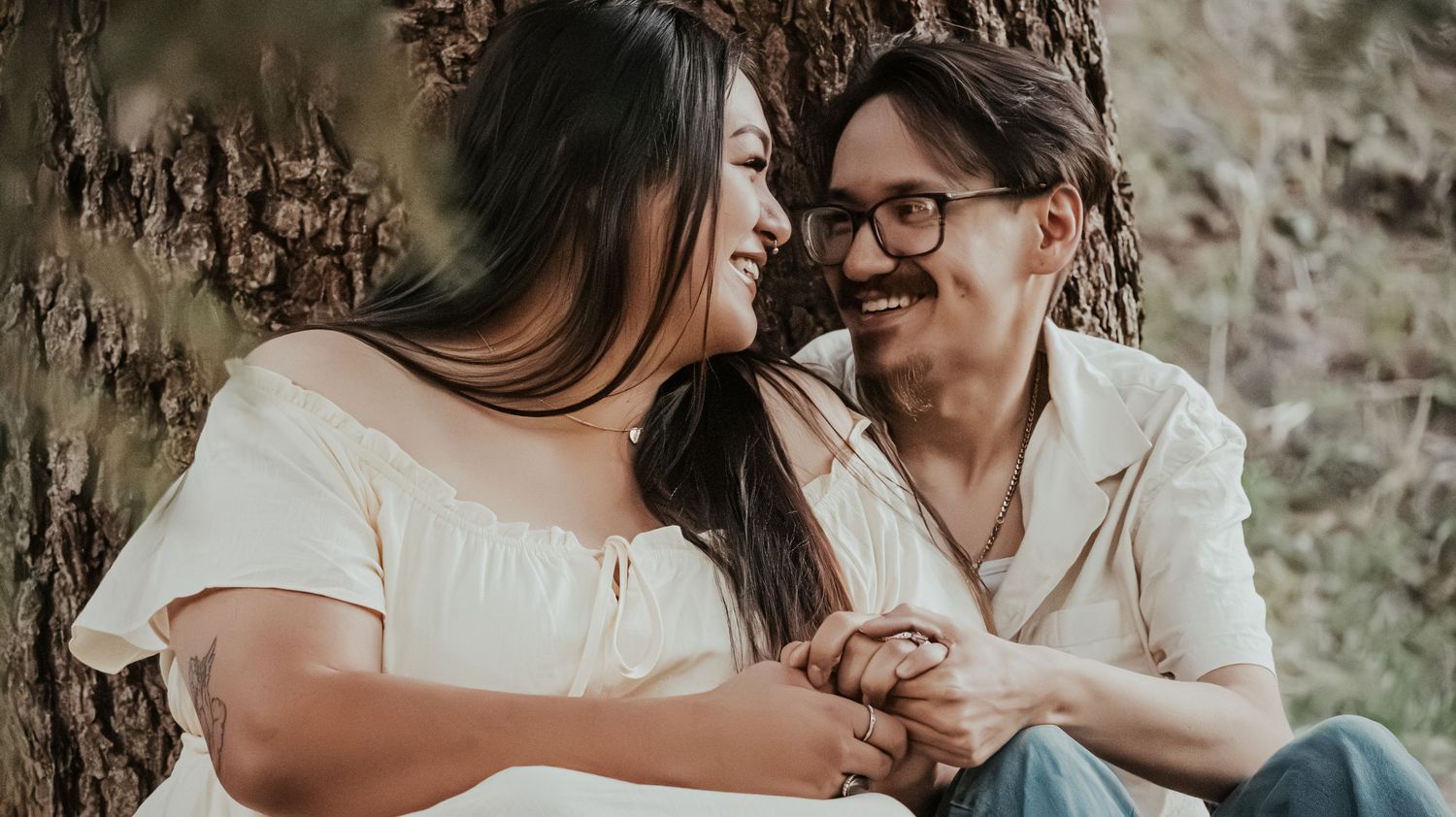A couple smiles and holds hands while sitting against a tree trunk outdoors.