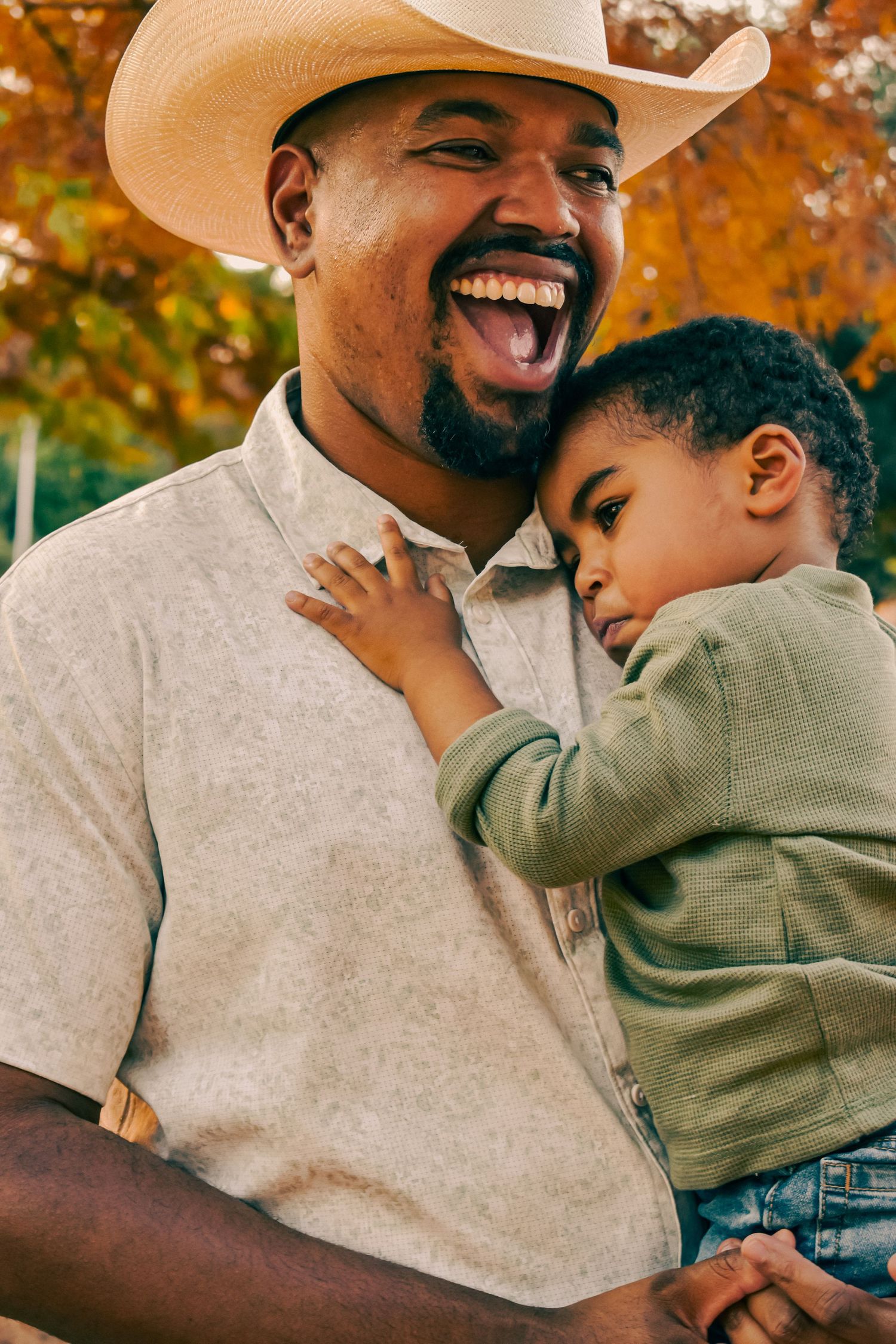 A joyful father in a cowboy hat laughs while holding his young child outdoors during autumn for a family session.