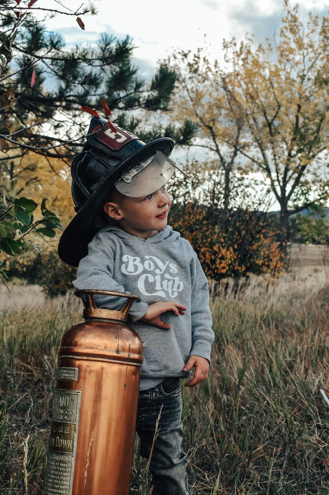 Toddler wearing firefighter helmet stands beside vintage copper fire extinguisher outdoors. Family Photographer in Pflugerville, Tx
