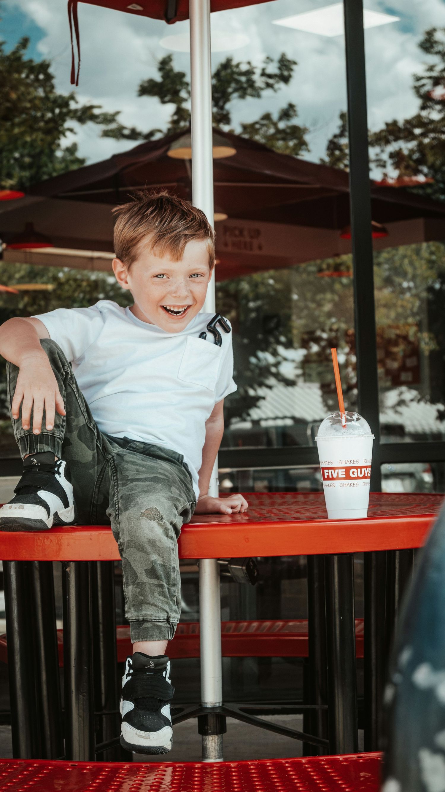 A happy boy sits on a red table at Five Guys restaurant, smiling with a milkshake nearby. Family Photographer in Pflugerville, Tx
