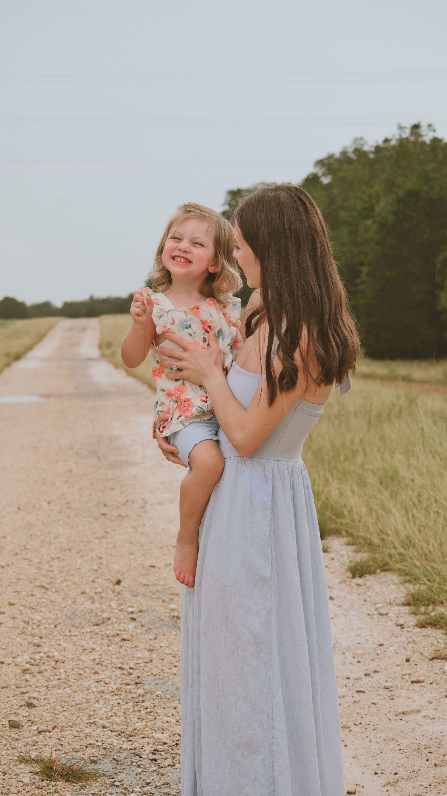 A young woman in a light blue maxi dress holds a smiling toddler girl on a sunny rural dirt road for a family session.