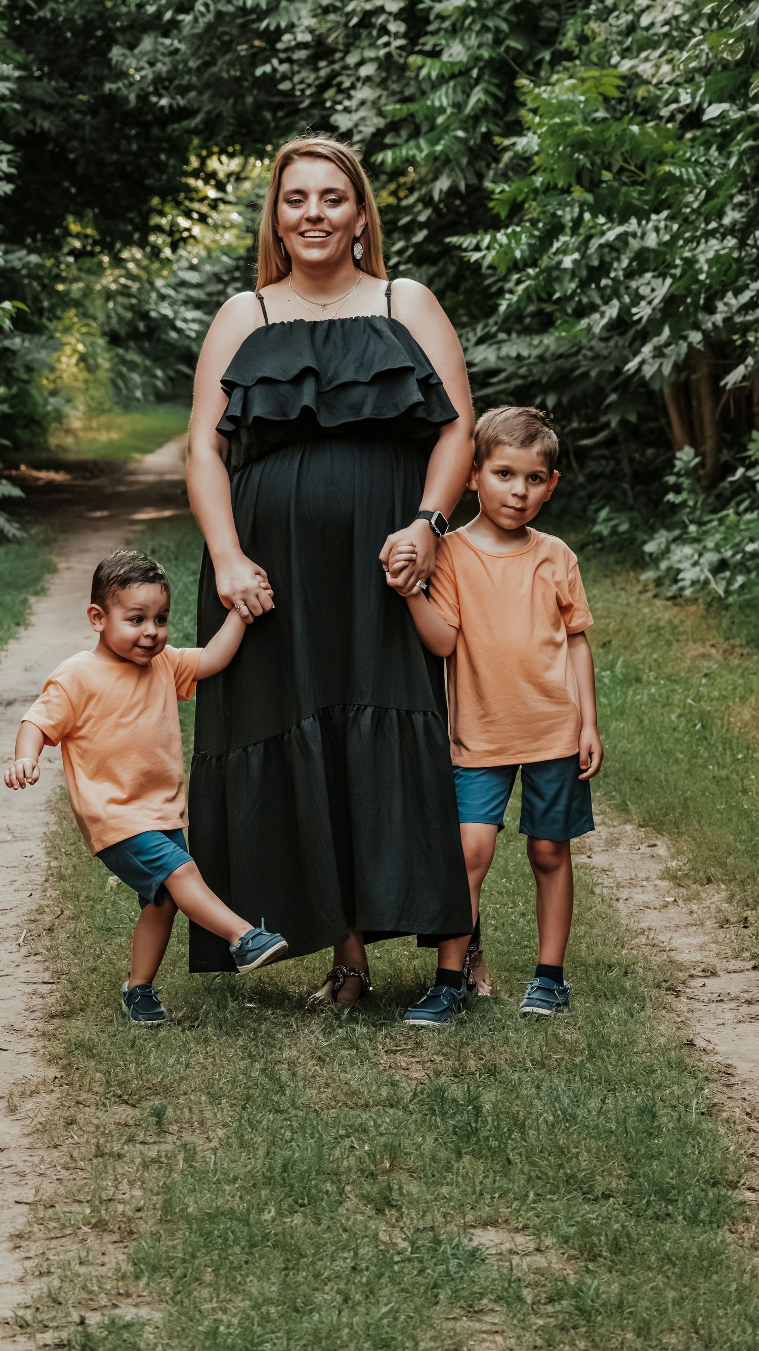Pregnant woman in black dress holding hands with two young sons in matching orange shirts on a park path.