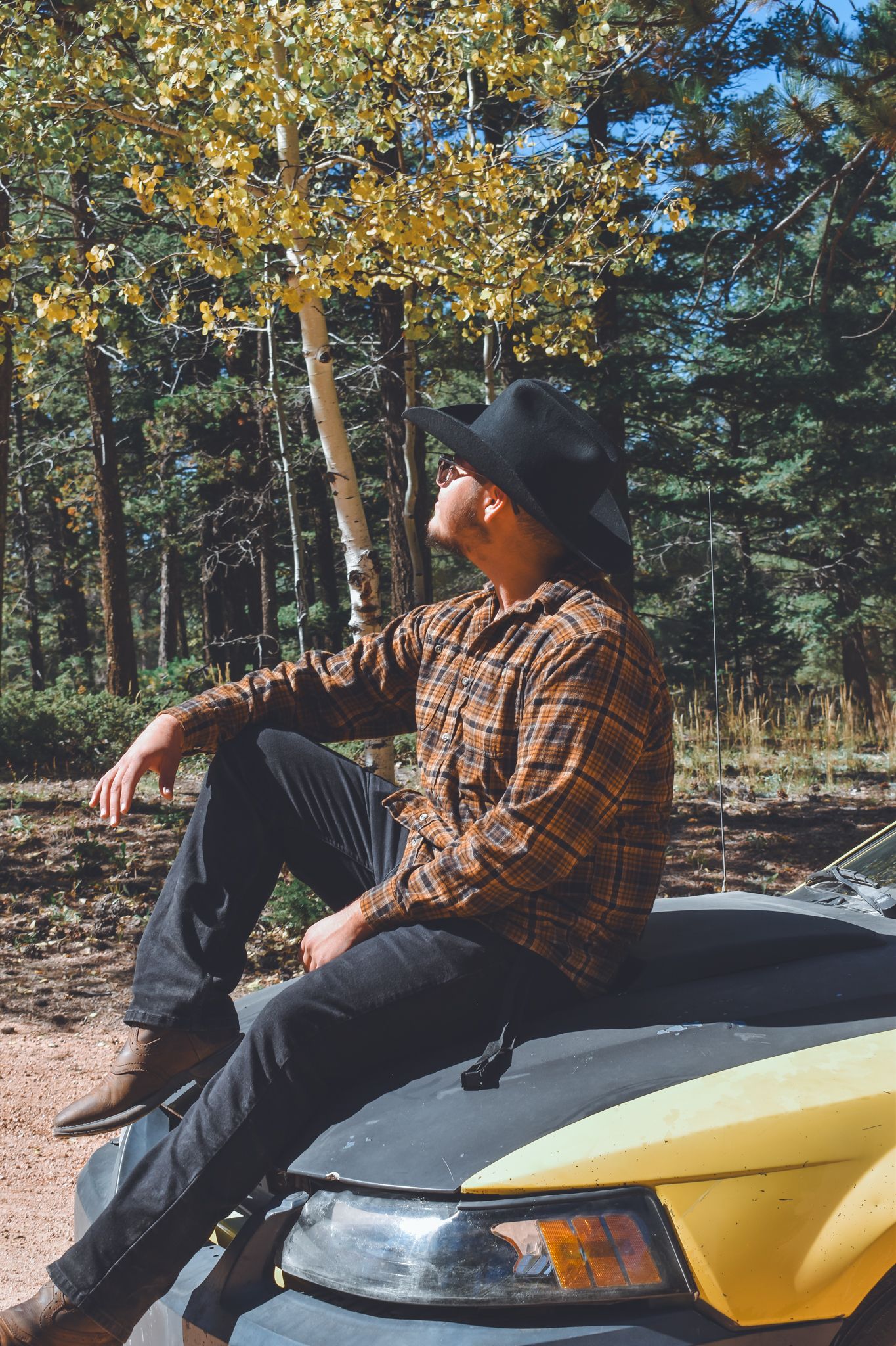 Man in cowboy hat sitting on hood of yellow car gazing up in an autumn forest for senior photos.