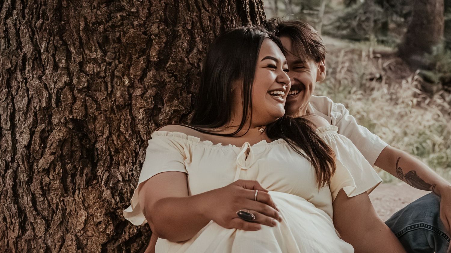 A joyful couple leans against a tree trunk, laughing together in a sunlit outdoor setting.